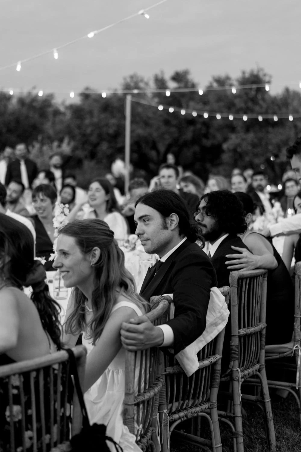 Una folla di persone sedute a un matrimonio all'aperto con luci pendenti, alcune sorridenti e in tight, in un ambiente naturale.