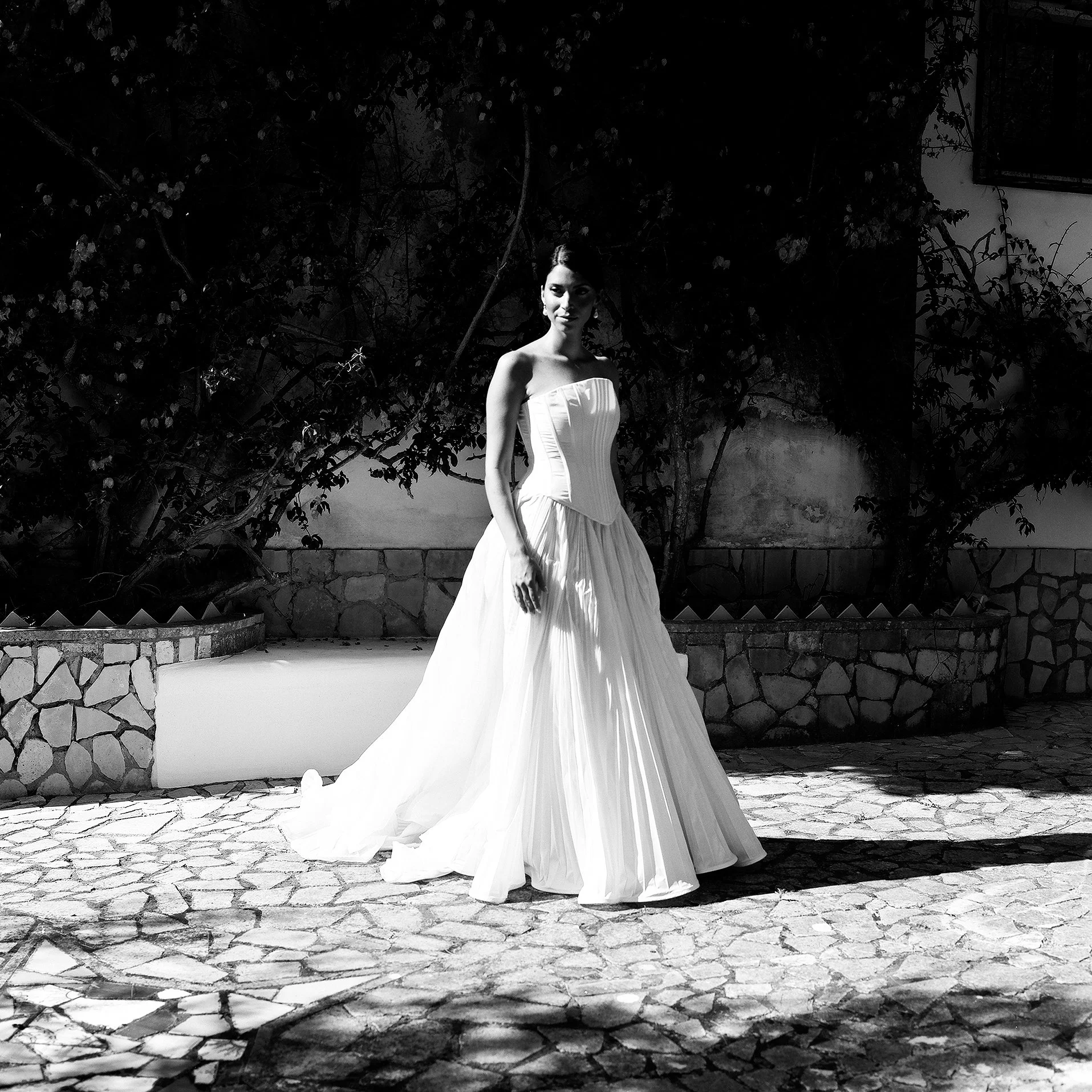 Donna in abito da sposa su un ciottolato, con piante e muro sullo sfondo.