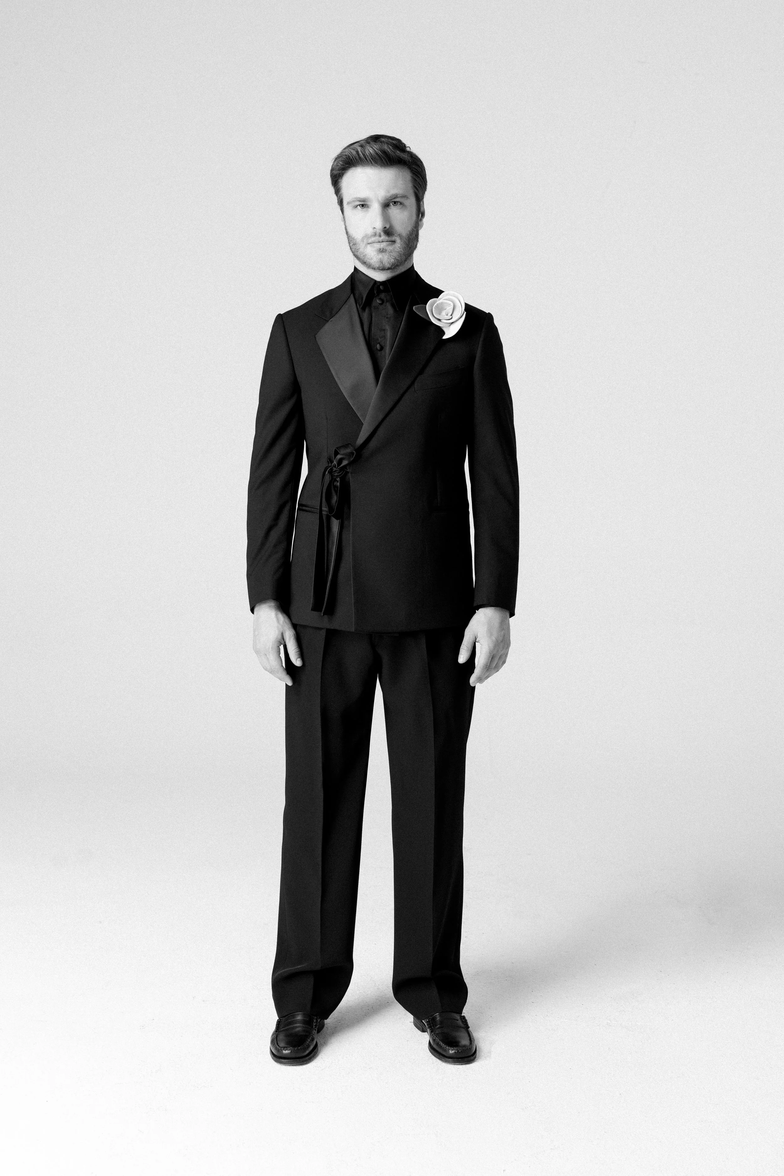 Uomo in abito nero con fiore bianco sulla giacca, in posa su sfondo bianco.