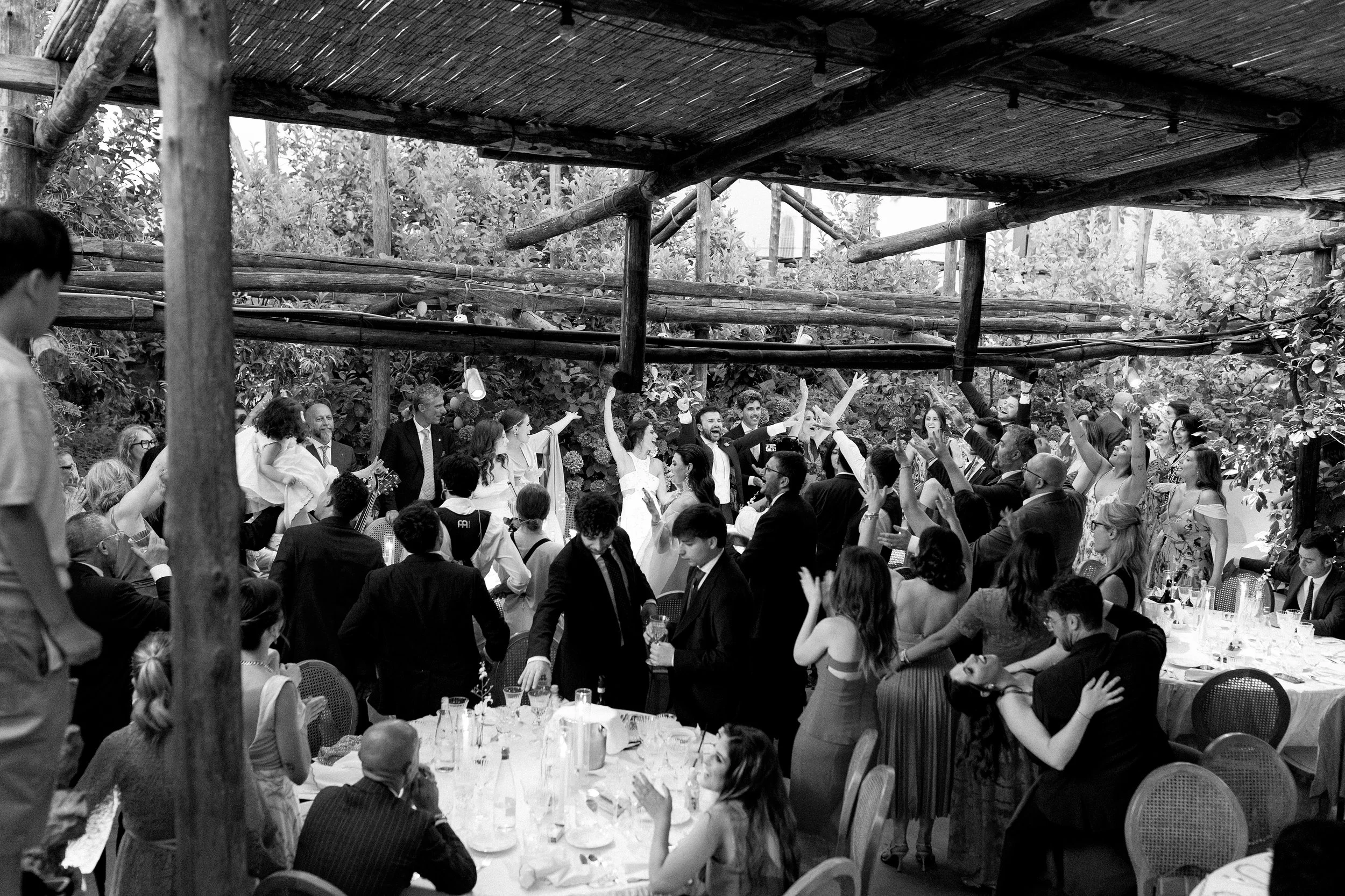 Una festa di matrimonio all'aperto con molti invitati che ballano e si divertono sotto una struttura di legno, ambientazione naturale con alberi sullo sfondo.