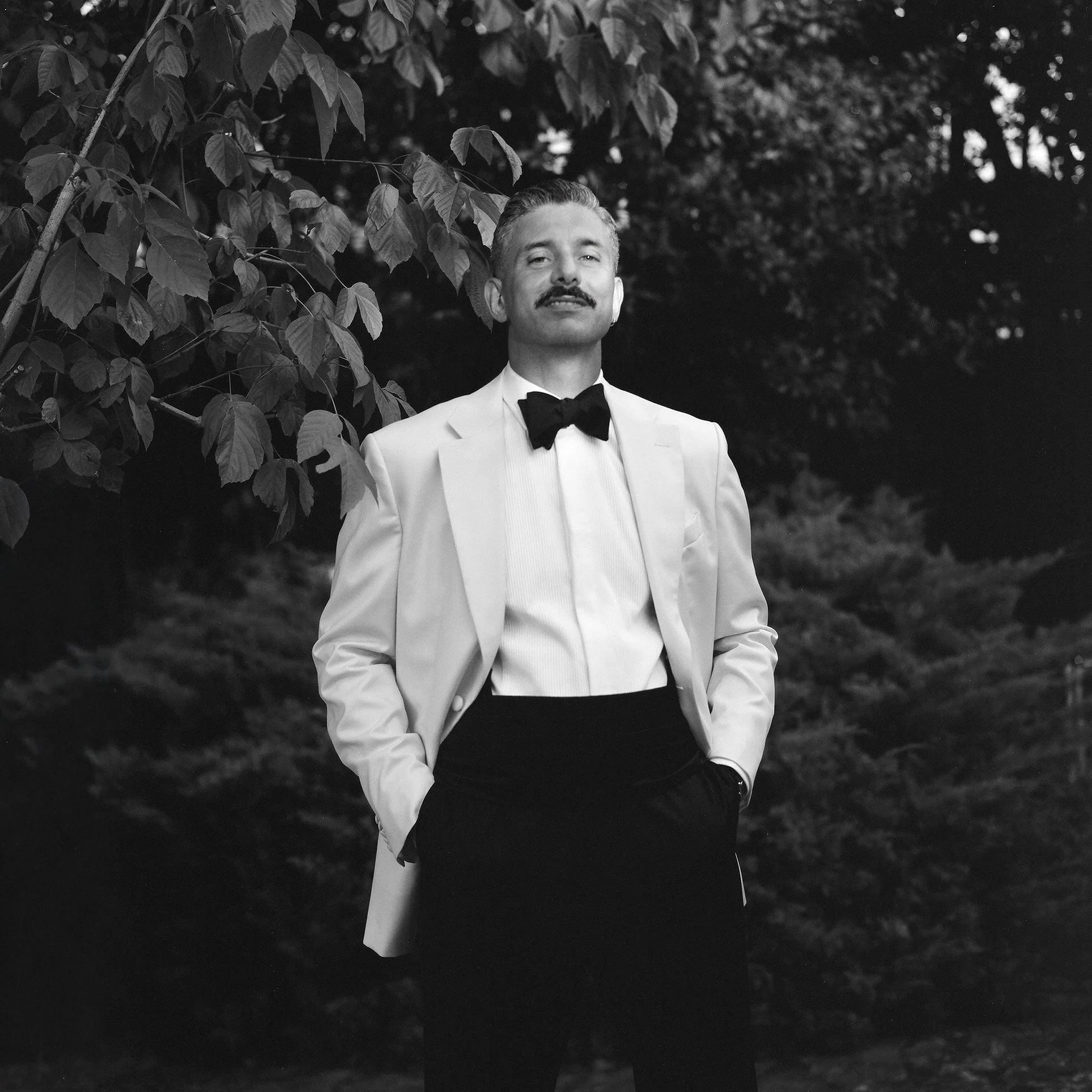 Uomo con baffi in abito bianco, papillon nero, posizionato di fronte a un albero, in un ambiente naturale.