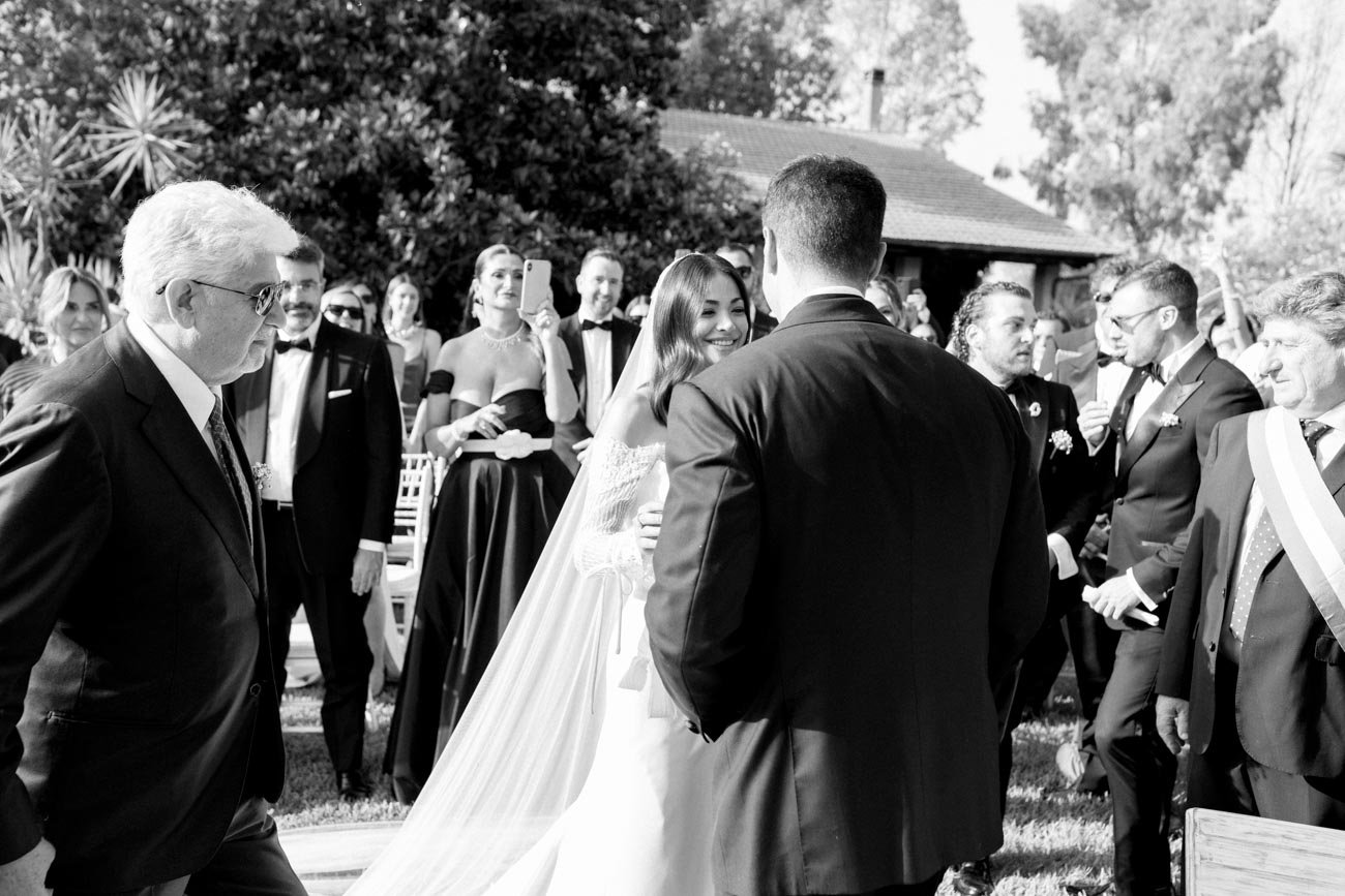 Photo in bianco e nero di un matrimonio all'aperto con persone vestite elegantemente che ridono e scattano foto, al centro una sposa e uno sposo che si guardano sorridendo.