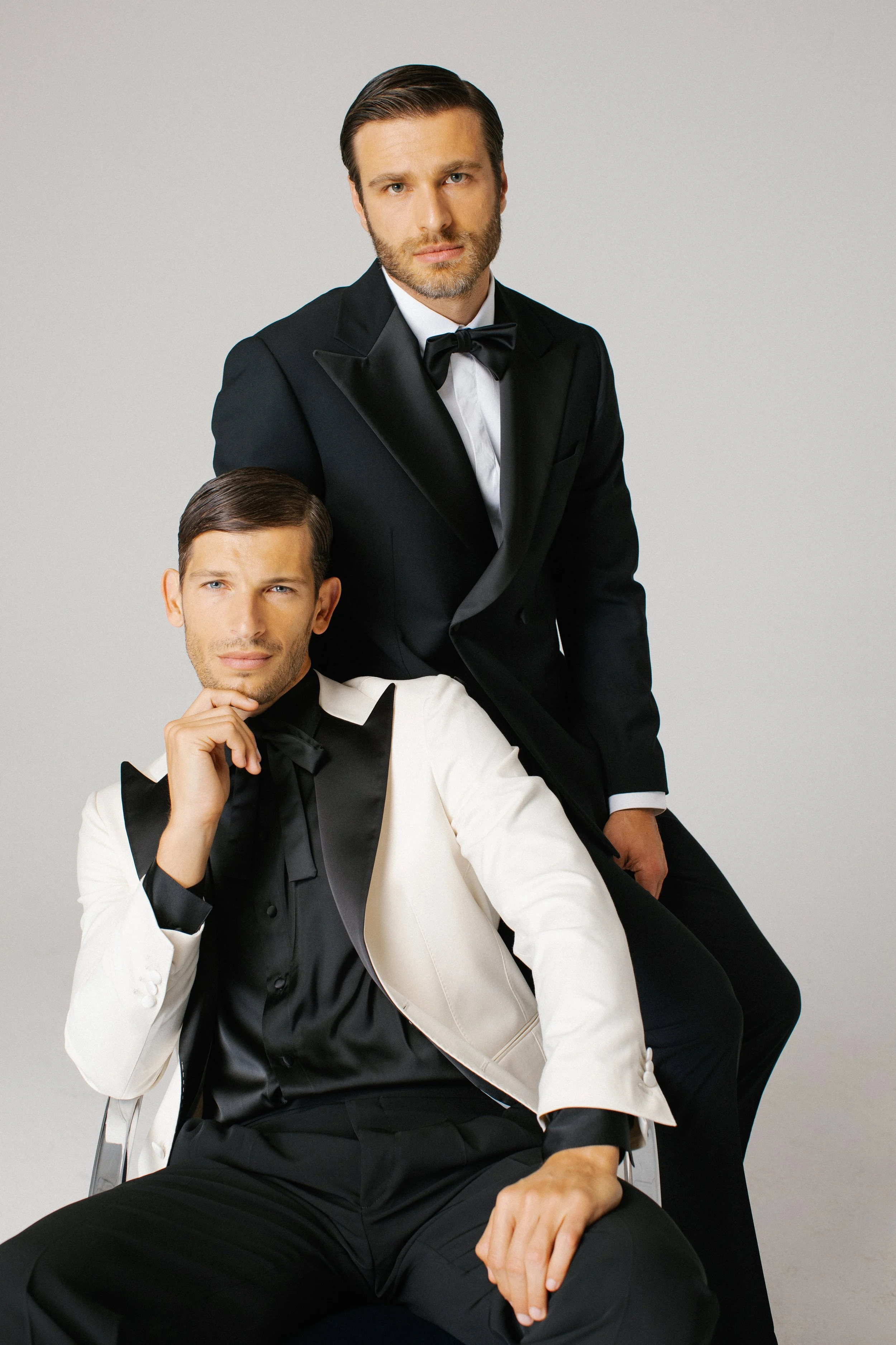 Due uomini vestiti elegantemente in smoking, uno seduto con una giacca bianca e uno in piedi con una giacca nera, entrambi con cravatte nere, posano su uno sfondo grigio chiaro.