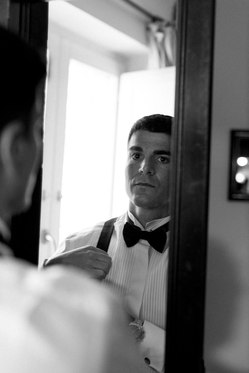 Uomo in abito da ore, con papillon, che si guarda nello specchio mentre si sistema le spalline della camicia