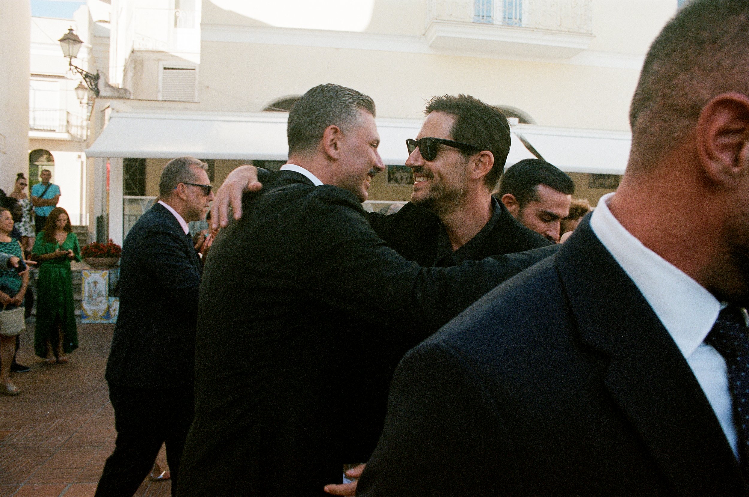 Uomini in abiti eleganti che si abbracciano e sorridono durante un evento sociale all'aperto, con alcuni altre persone in sottofondo e un edificio bianco sullo sfondo.