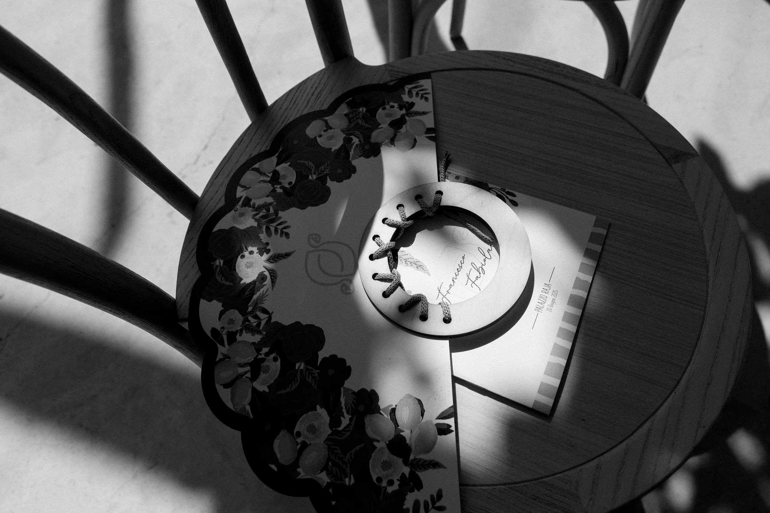 Un cappello di paglia con decorazioni floreali e un berretto con lacci, posato su una sedia di legno, in una luce che crea ombre.