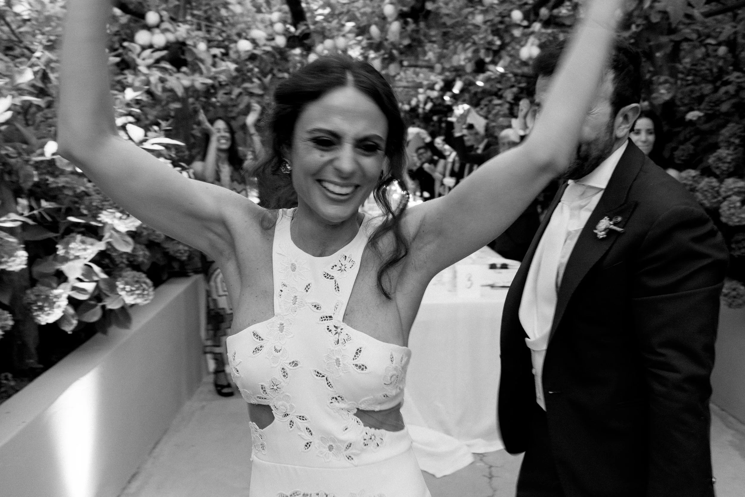 Una donna felice con un vestito bianco ricamato, con le braccia sollevate mentre balla o festeggia, in un ambiente decorato con fiori, vicino a un uomo in abito nero, probabilmente durante un matrimonio o una celebrazione.