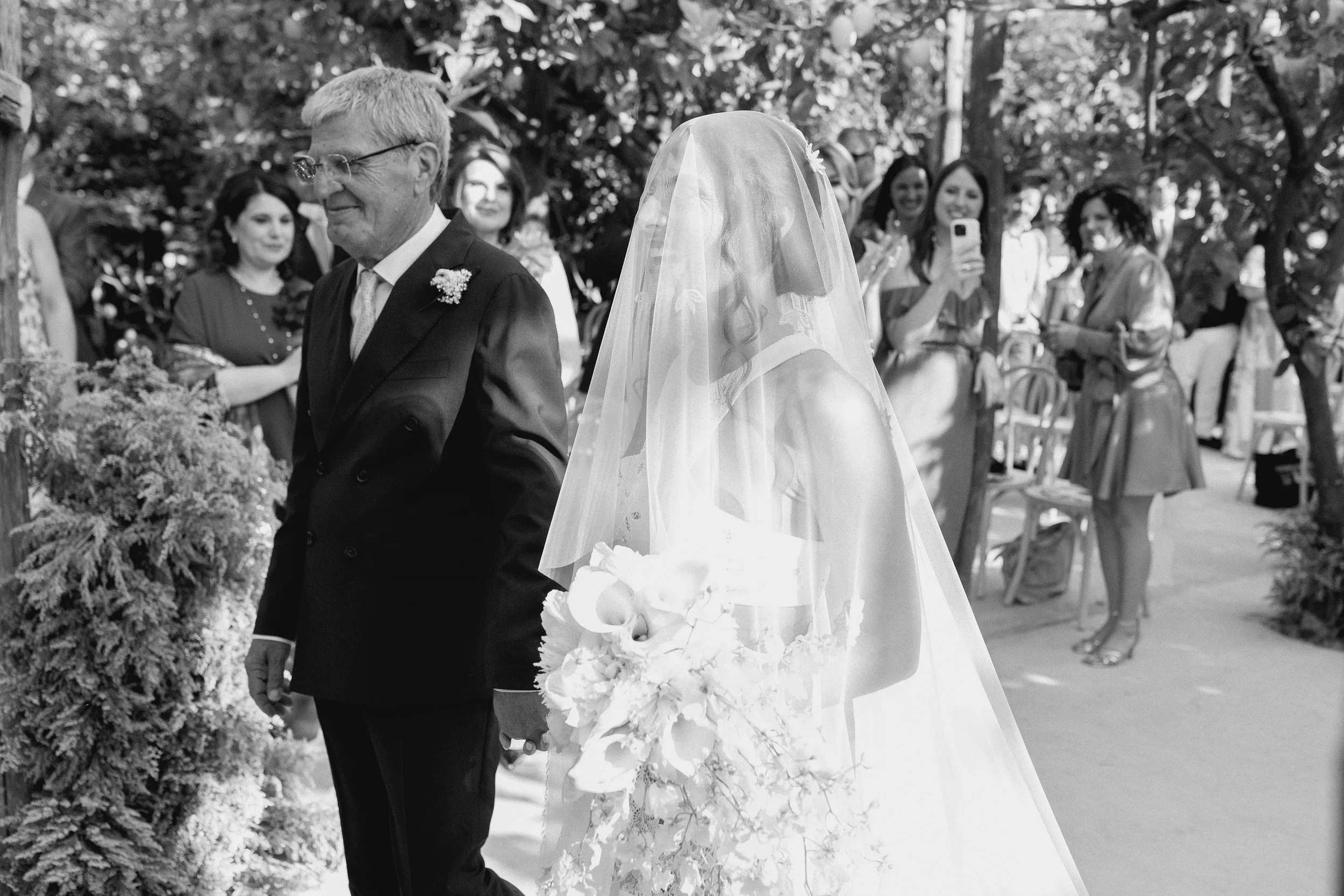 Una coppia di sposi, la sposa con un abito bianco e un velo, tiene un bouquet di fiori, in piedi accanto a un uomo anziano in abito scuro, circondati da invitati che assistono alla cerimonia all'aperto.