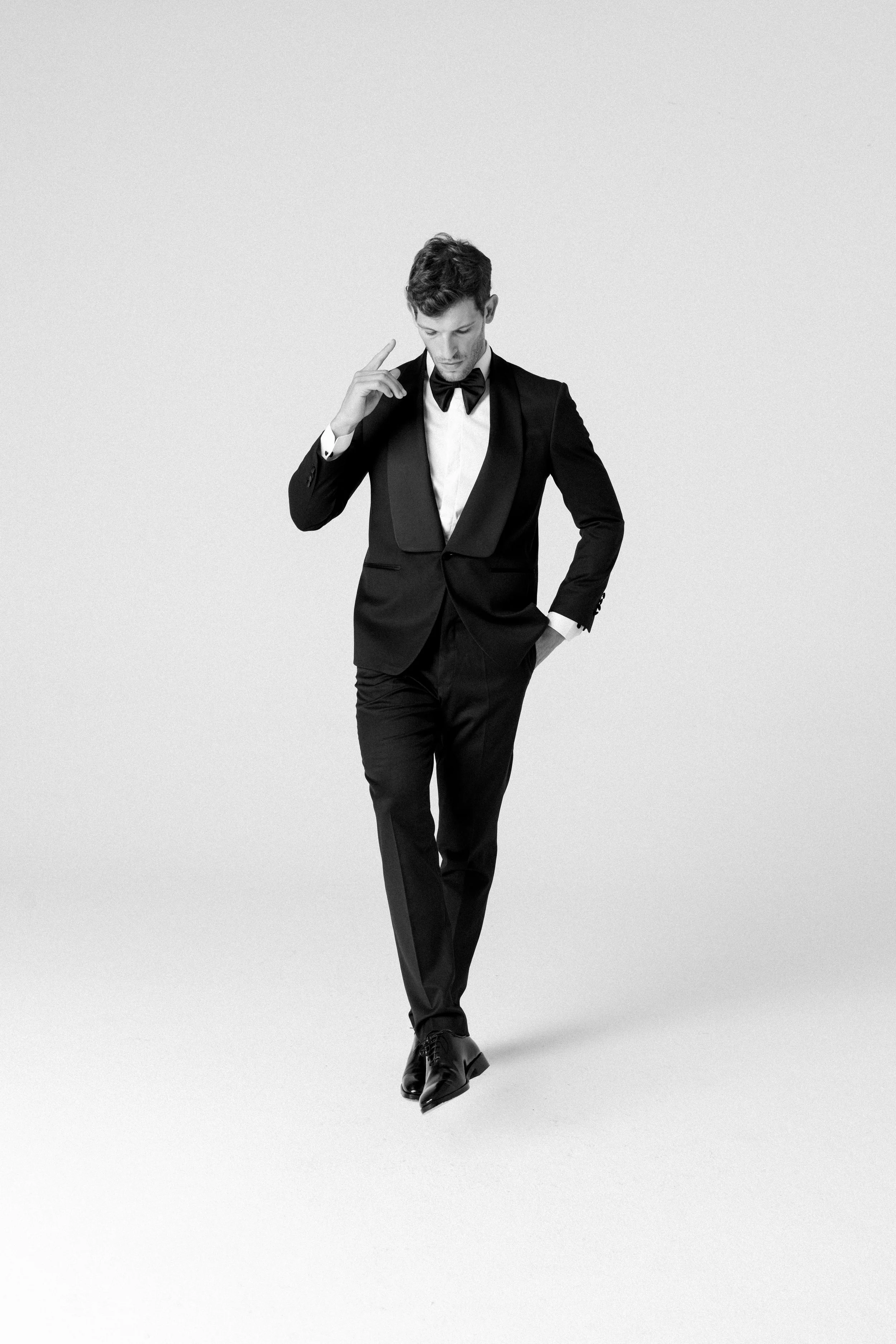 Uomo elegante in abito nero con papillon, che cammina e si manca di una mano, in uno sfondo bianco.