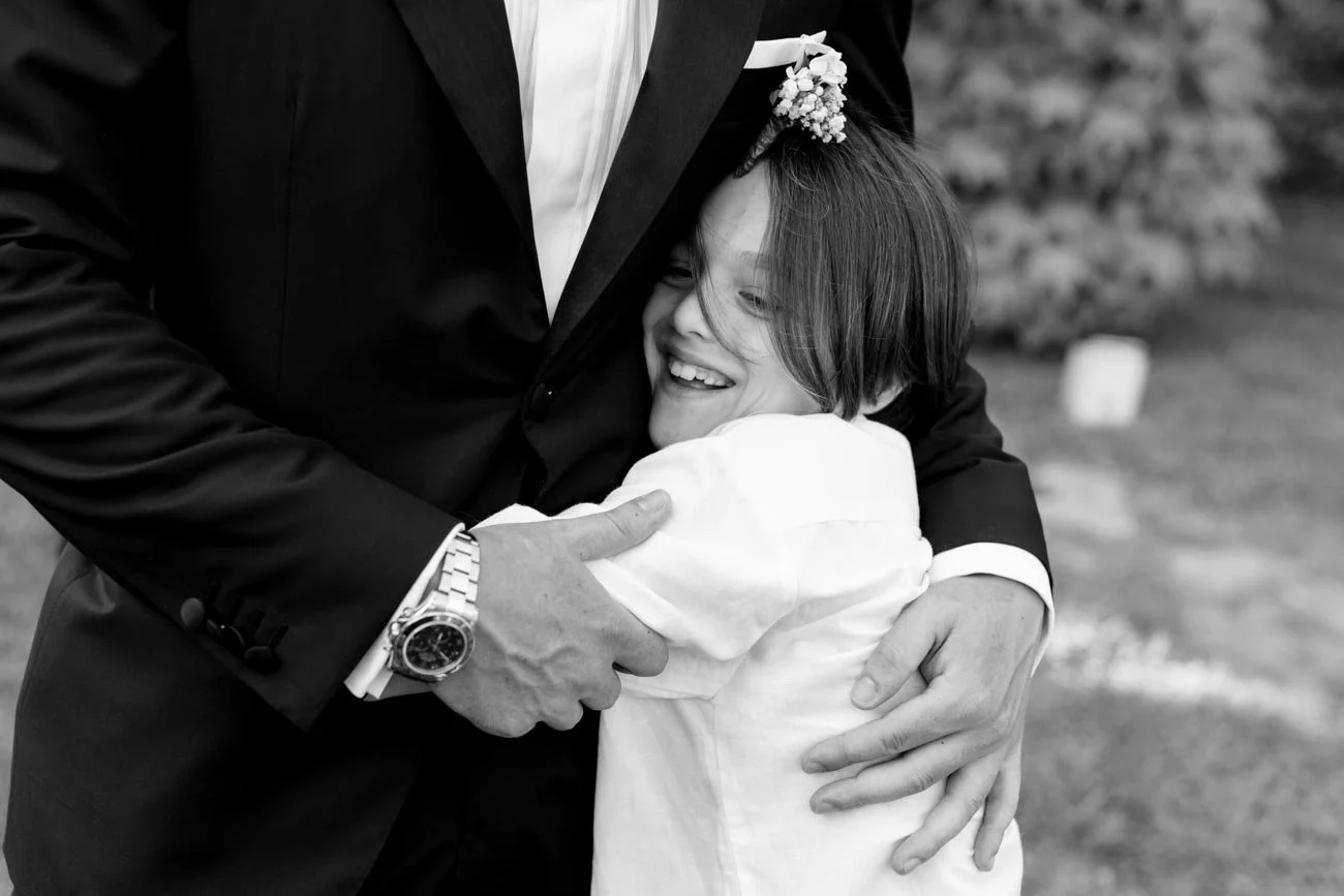 Due persone si abbracciano, una donna sorridente con un fiore tra i capelli e una persona con abbigliamento formale, in un'ambientazione all'aperto.