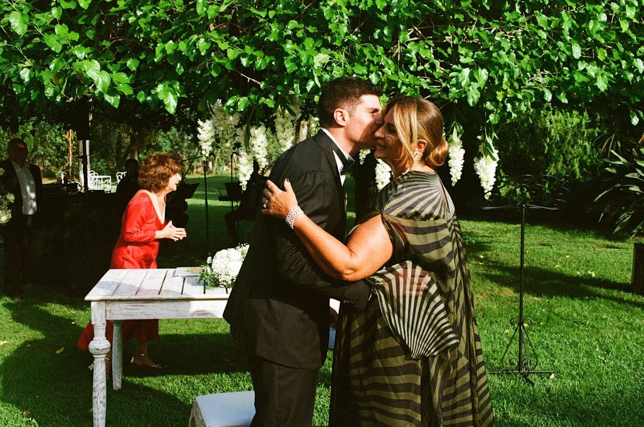 Una coppia si abbraccia e si bacia durante una cerimonia all'aperto, con un tavolo di decorazioni floreali e altre persone sullo sfondo, sotto un albero verde.
