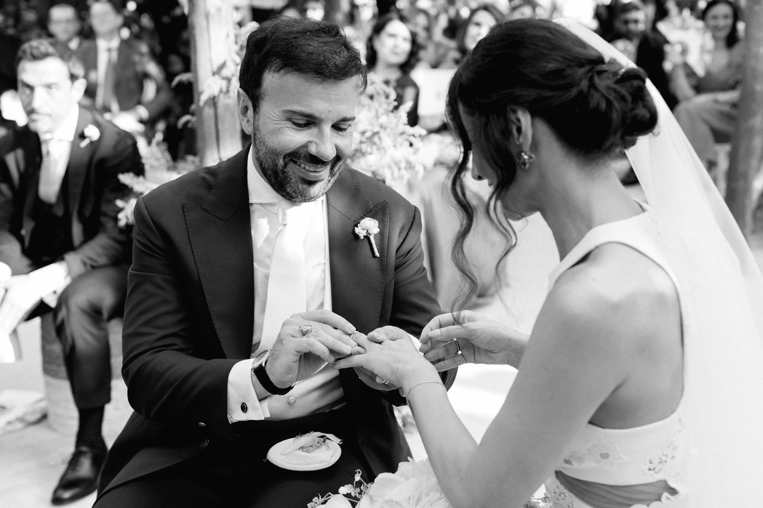 Una coppia si sposa, con l'uomo che mette l'anello alla sposa in un matrimonio all'aperto, circondati da invitati.