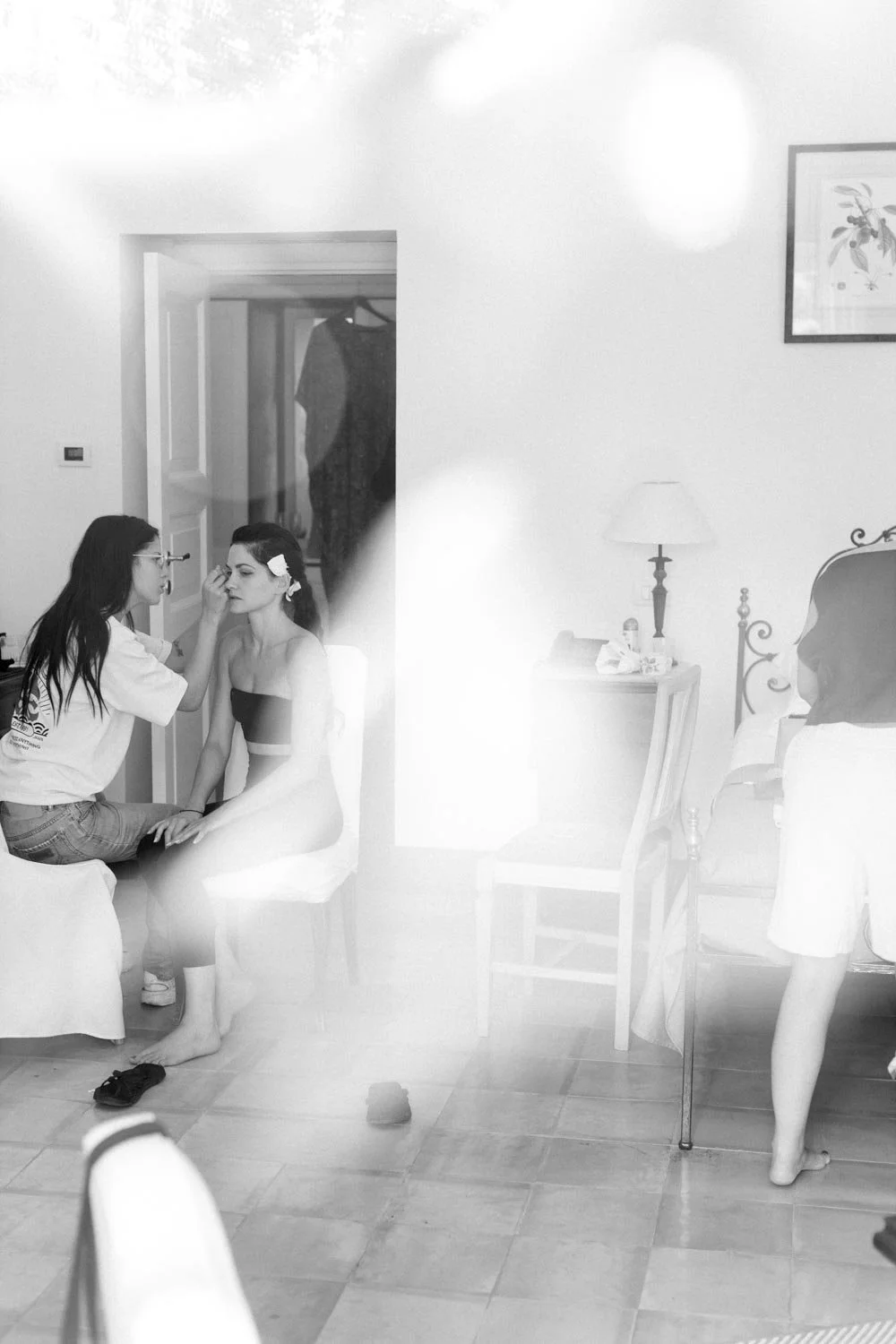 Una donna si sta facendo truccare da una truccatrice seduta su una sedia in una stanza. Un'ammagine in bianco e nero di un ambiente interno con mobili e quadri alle pareti.