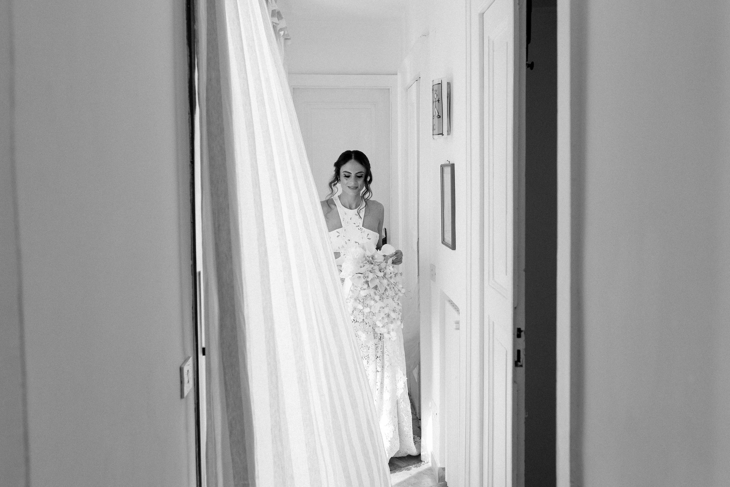 Una donna sorridente in abito da sposa con bouquet di fiori si affaccia da una porta.