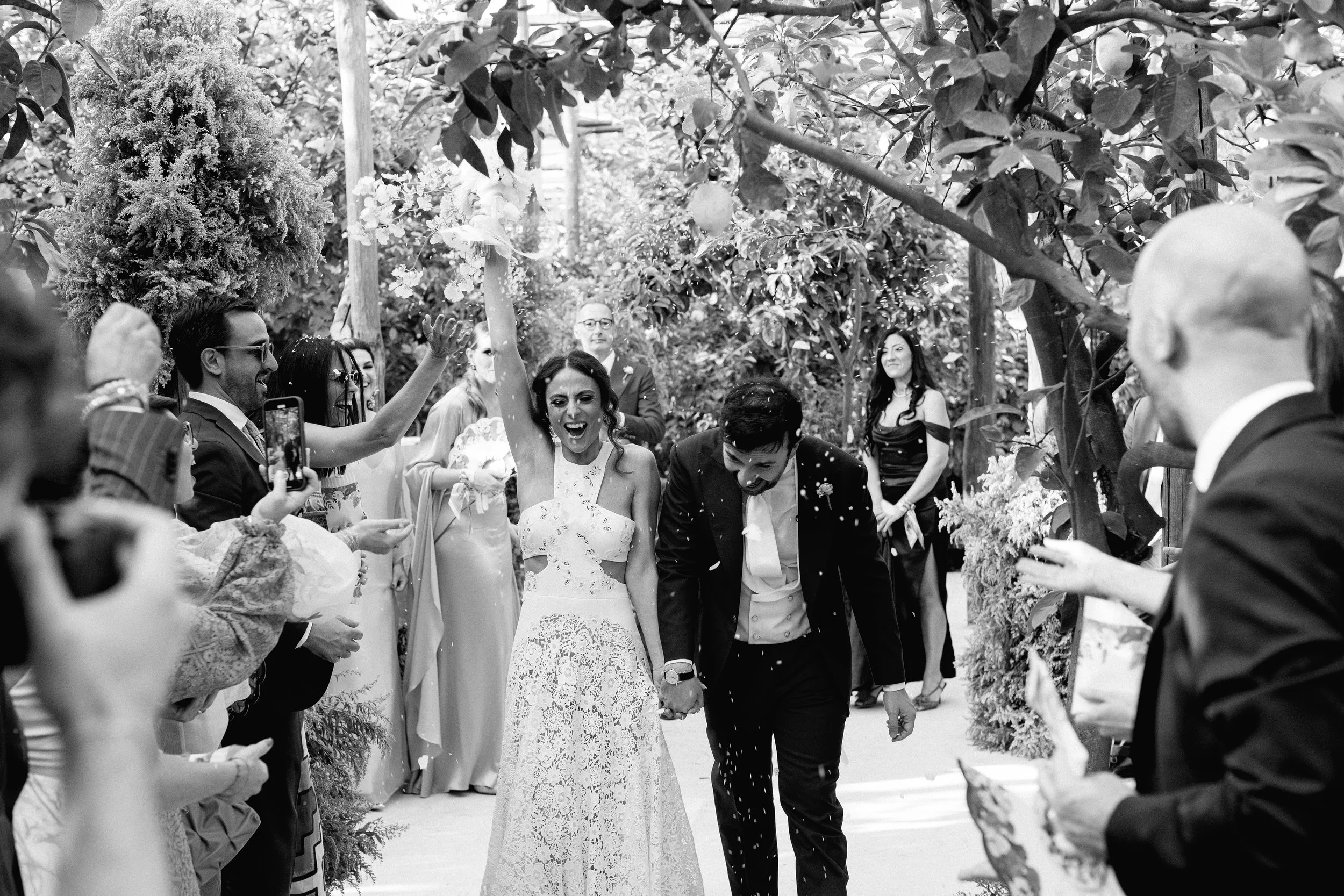 La coppia di sposi festeggia dopo il matrimonio, circondata da amici e familiari in un giardino, con il cielo e gli alberi sullo sfondo.