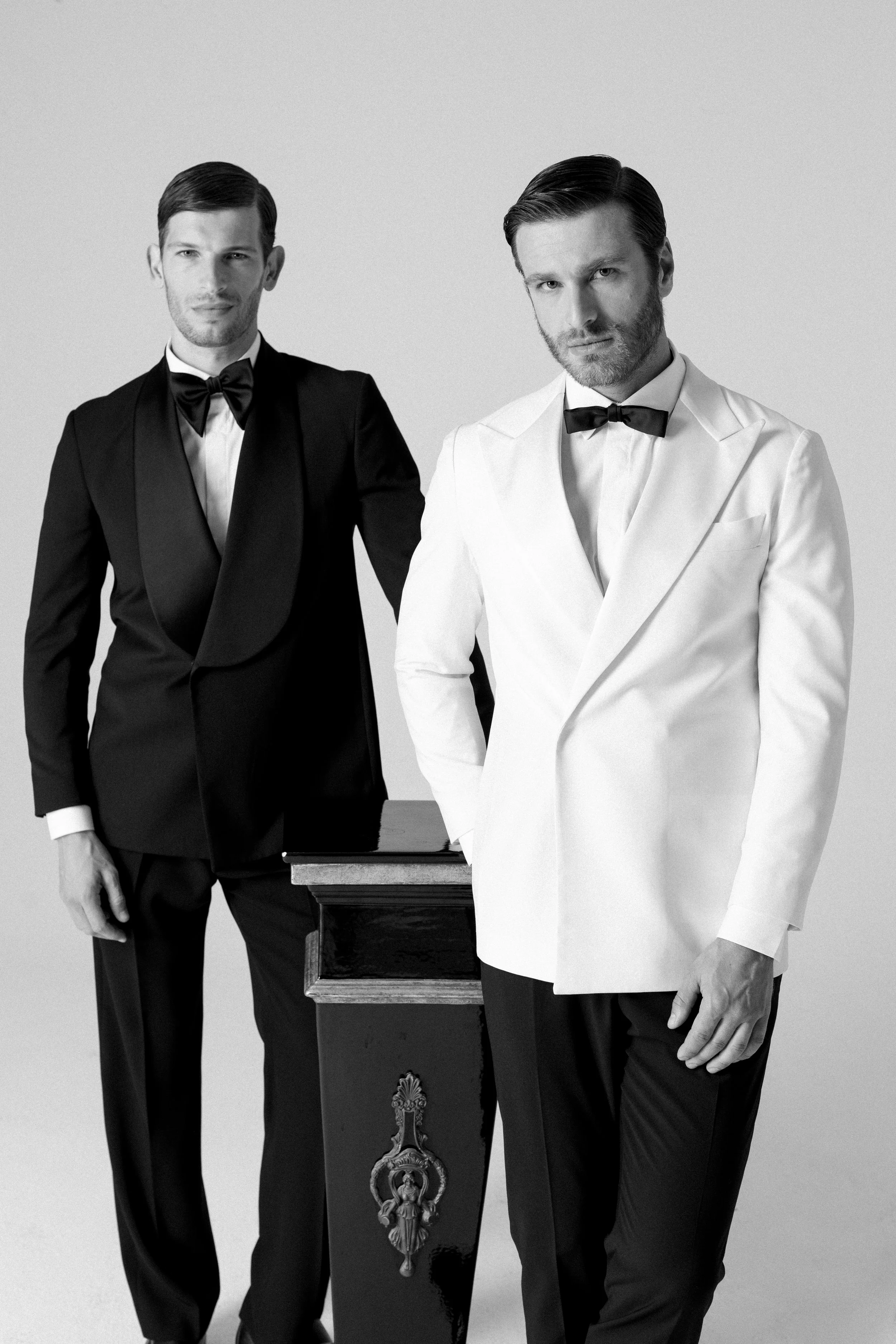Due uomini in abiti eleganti, uno in smoking nero e l'altro in smoking bianco, posano davanti a un tavolo di legno decorato con un emblema.