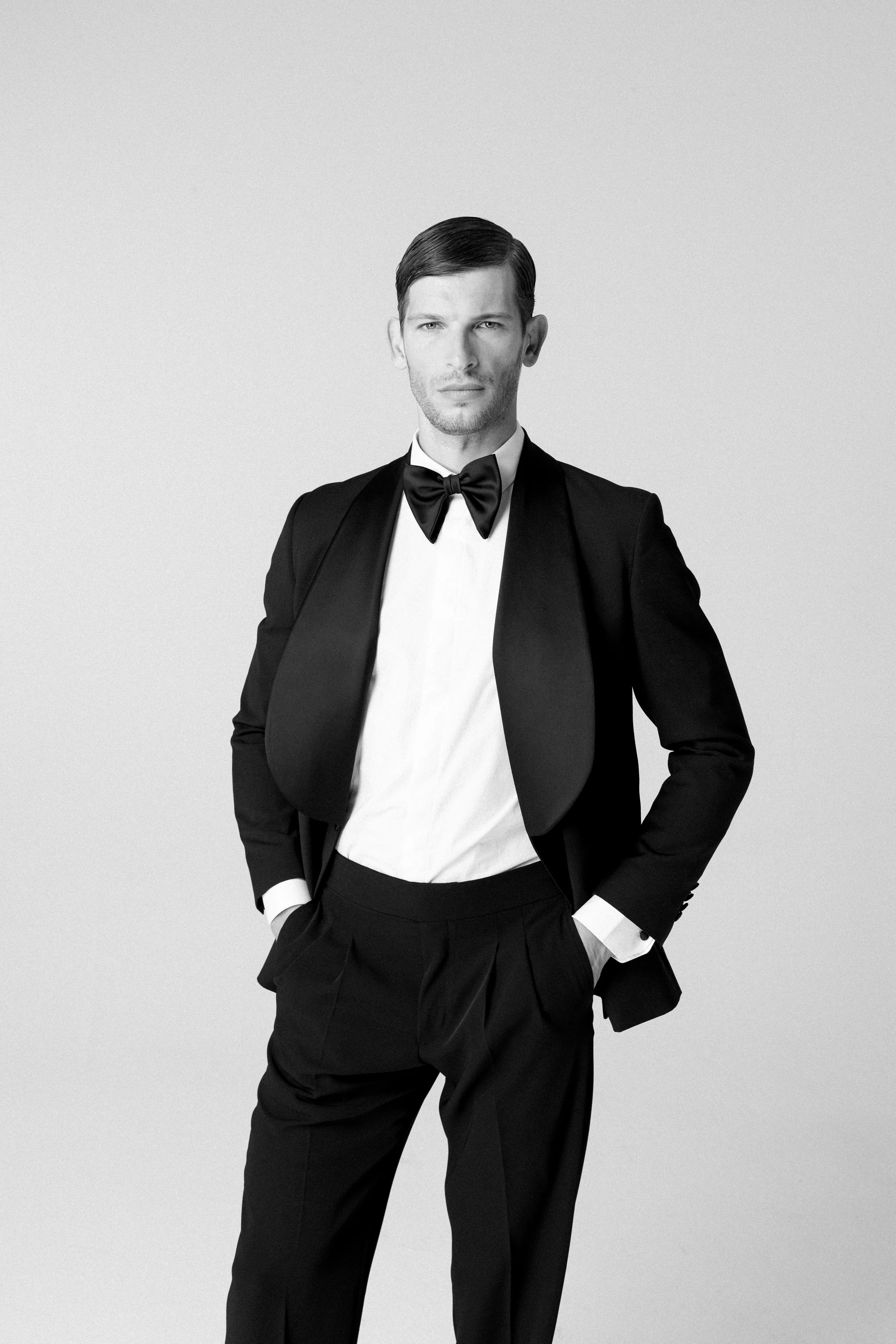 Uomo vestito in smoking con papillon nero, posa con le mani in tasca, sfondo chiaro