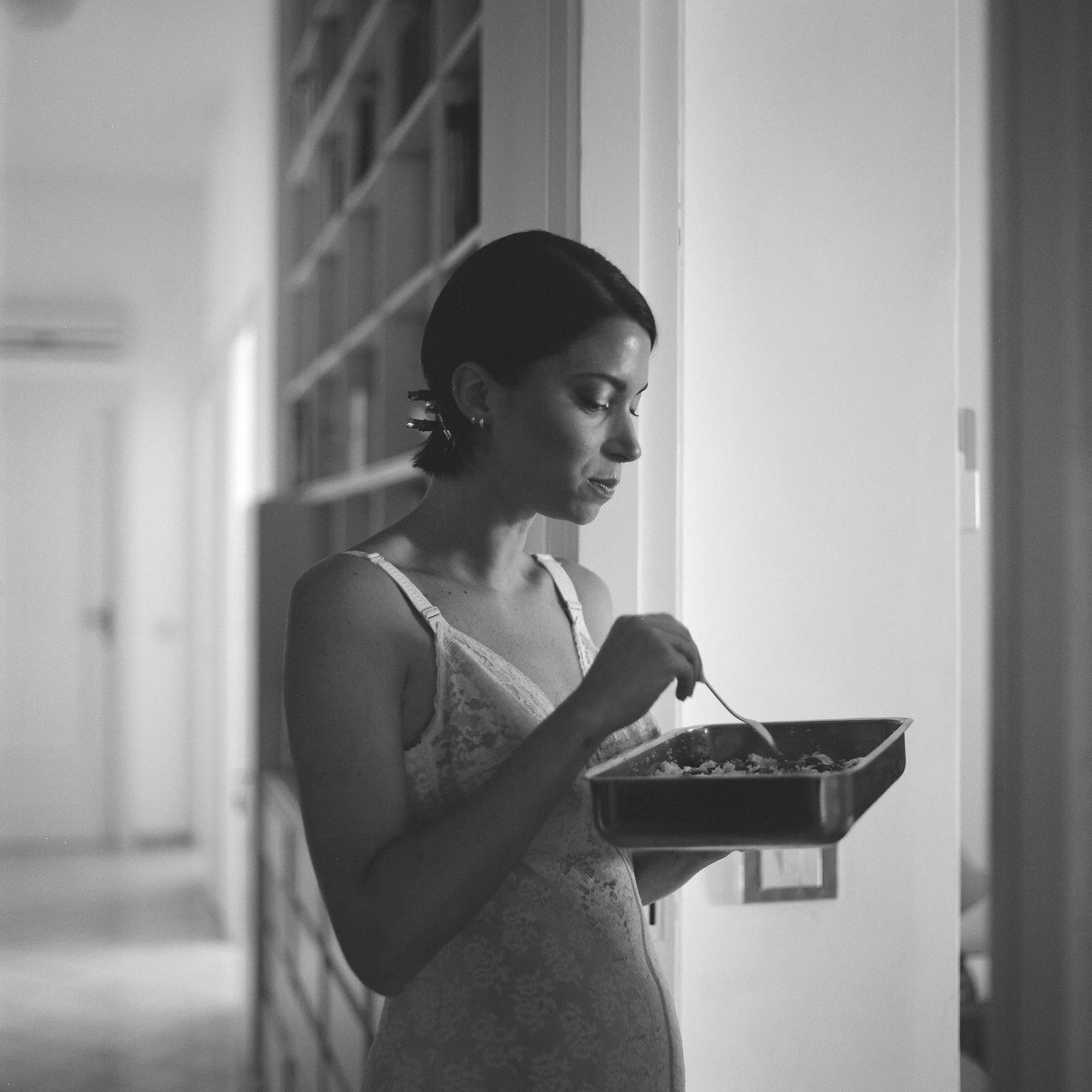 Donna con capelli scuri e occhi chiusi, indossa un vestito di pizzo, mentre mangia da un piatto con un cucchiaio, in un ambiente domestico con scaffali pieni di libri sullo sfondo.