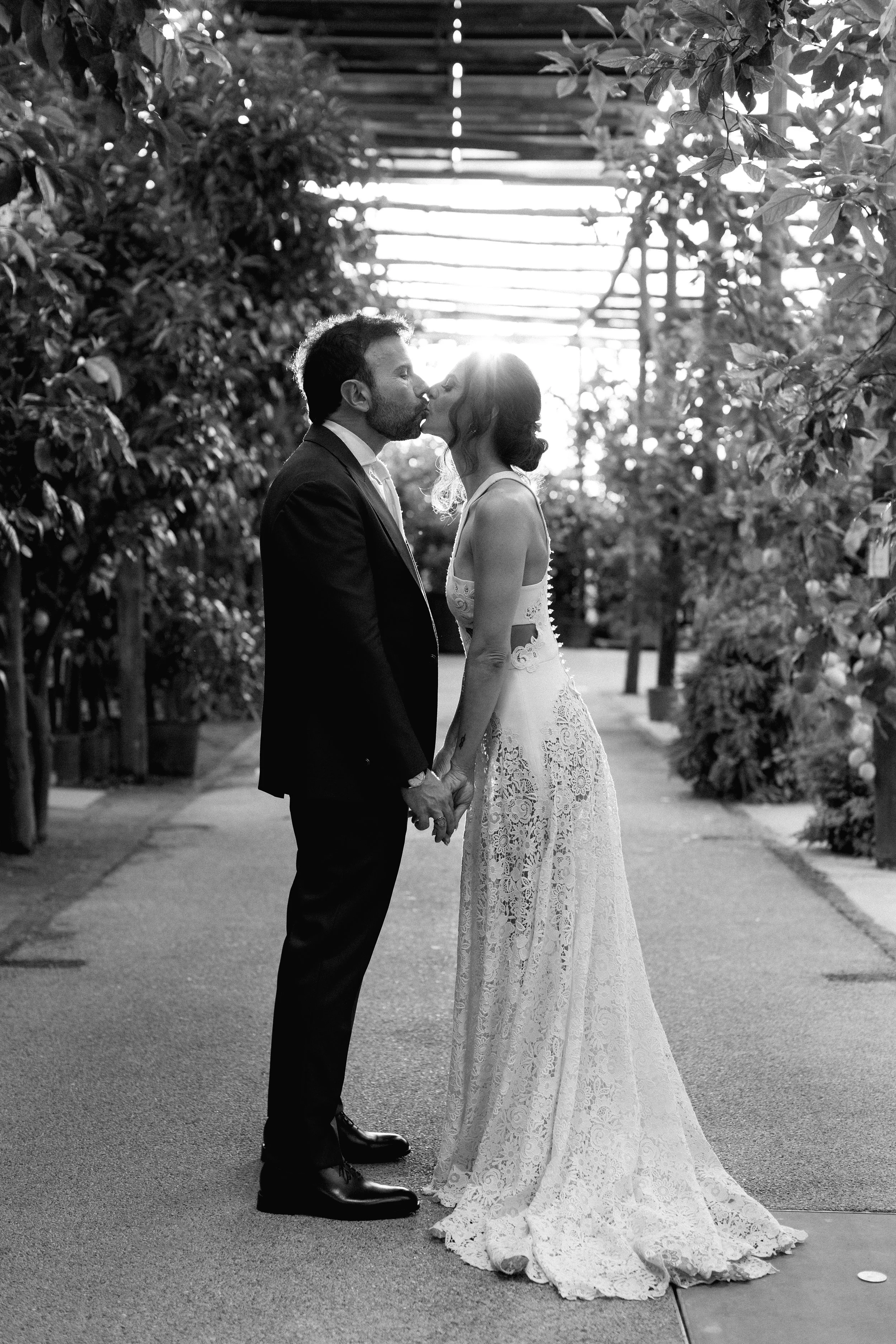 Una coppia di sposi si bacia in un luogo all'aperto, si tengono per mano, con il sole che splende dietro di loro, in un ambiente naturale.