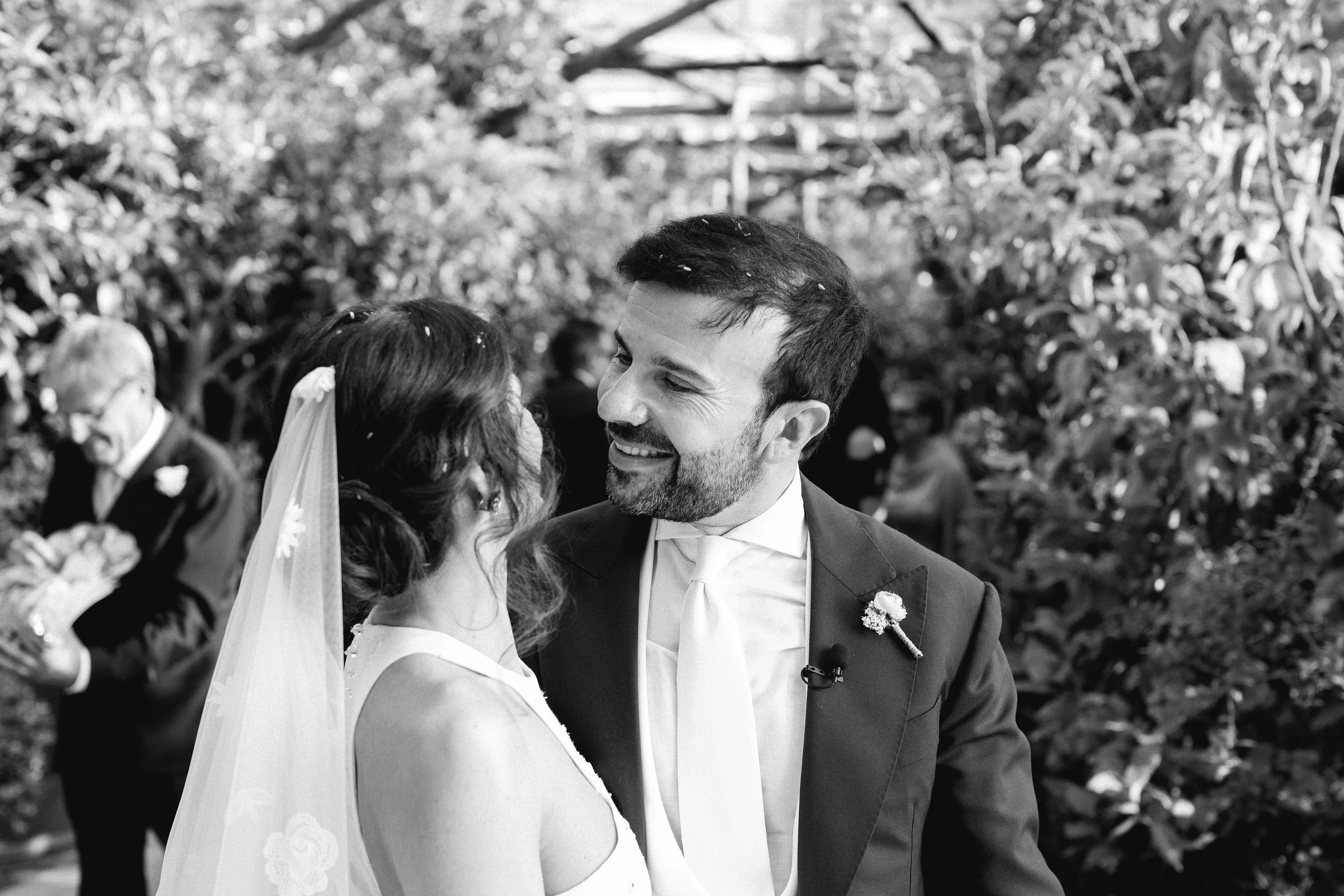 Coppia di sposi che si guarda negli occhi durante il matrimonio in un giardino, con altre persone sullo sfondo.