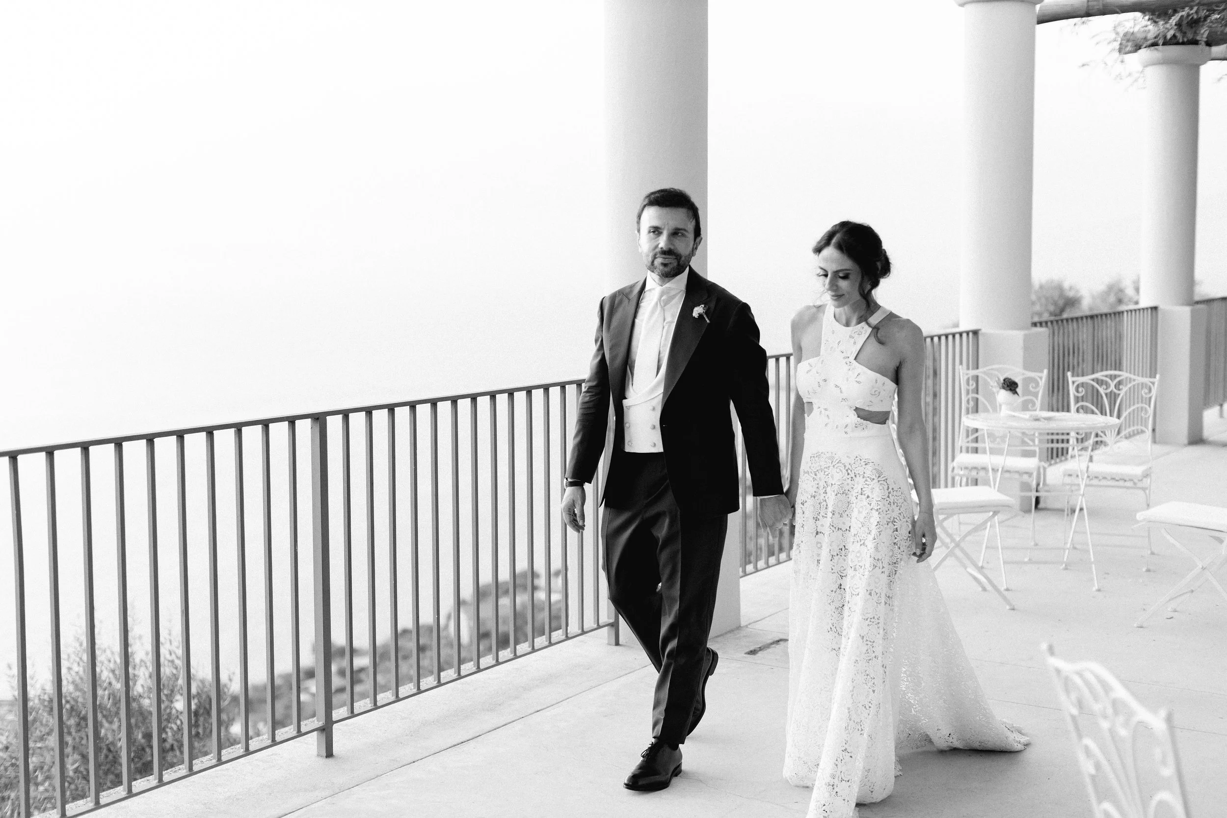 Una coppia di sposi in abiti da matrimonio cammina su un balcone aperto con sedie e tavoli in ferro battuto.