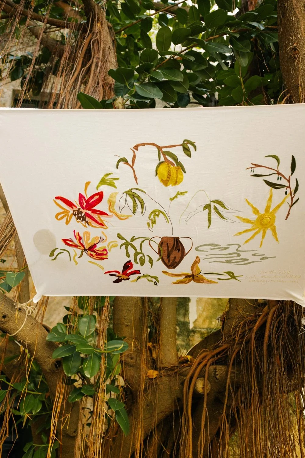 Tessuto bianco con disegni di fiori e piante, appeso tra rami di un albero con foglie verdi e radici visibili.
