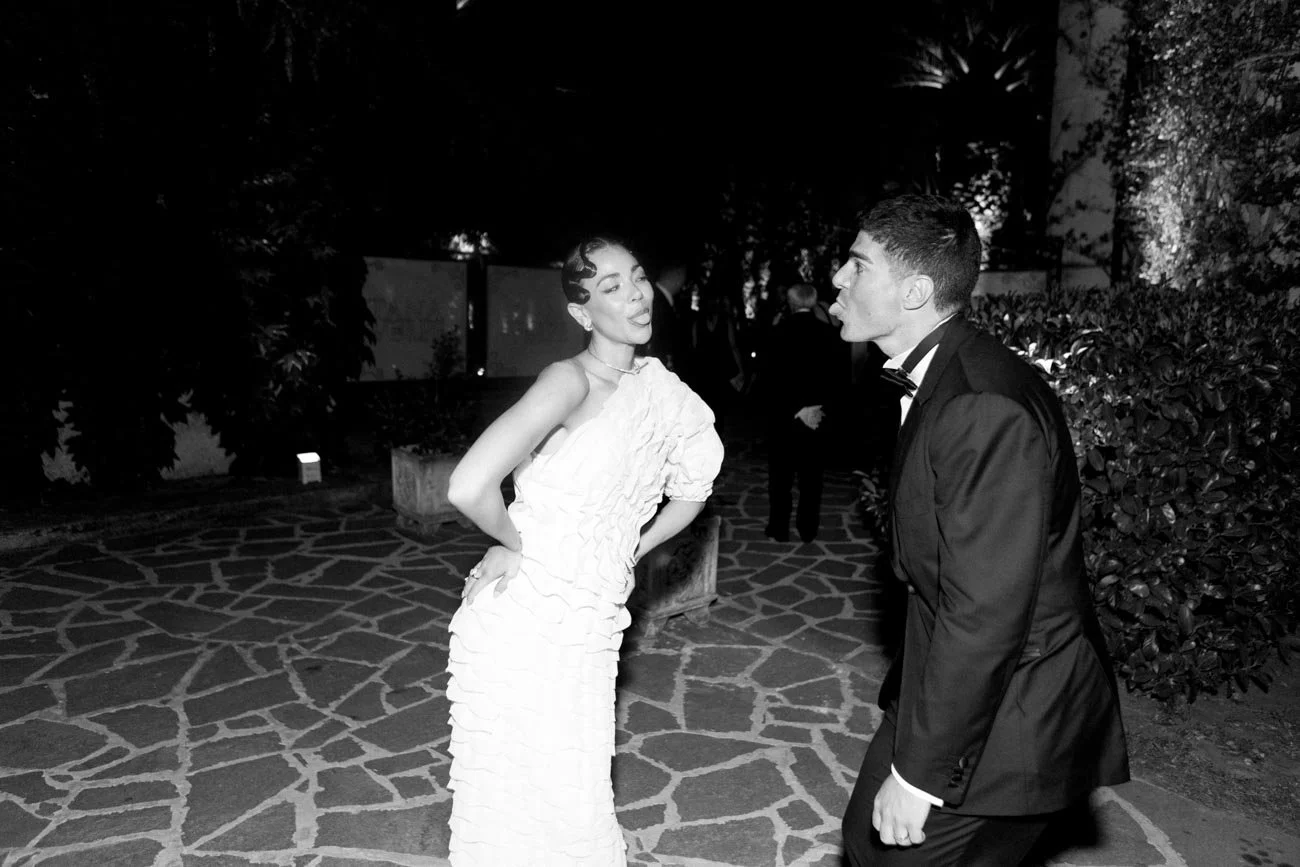 Due persone in abiti eleganti sembrano discutere o litigare all'aperto di notte, con alberi e piante sullo sfondo.