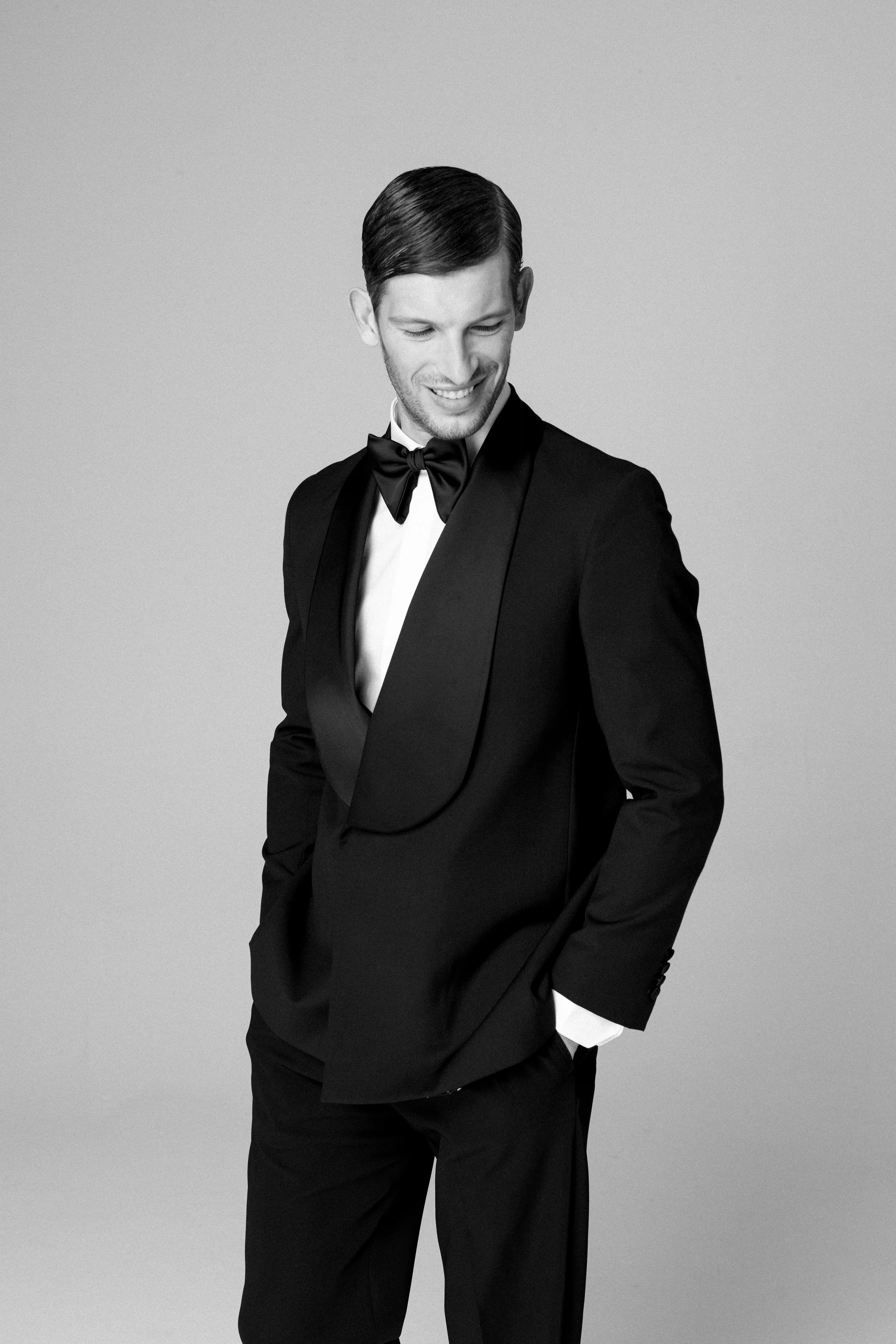 Uomo sorridente vestito in smoking nero con papillon, posa in modo elegante contro sfondo semplice.