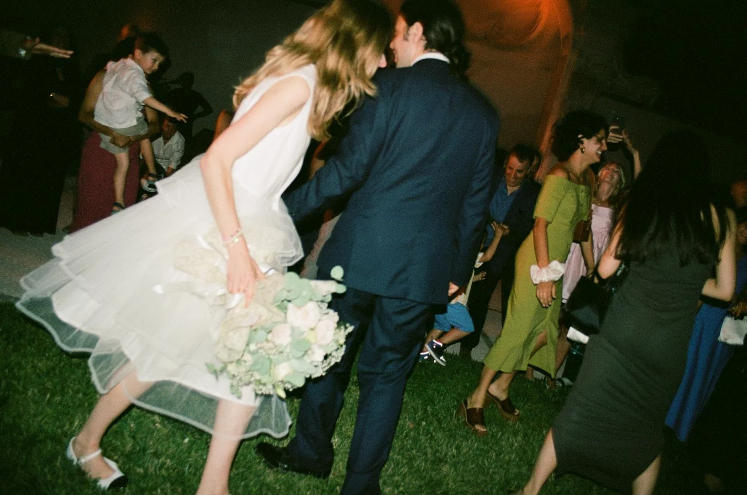 Persone che ballano e si divertono in un ambiente festivo, con un uomo in giacca scura e una donna in vestito bianco che tengono in mano un mazzo di fiori.