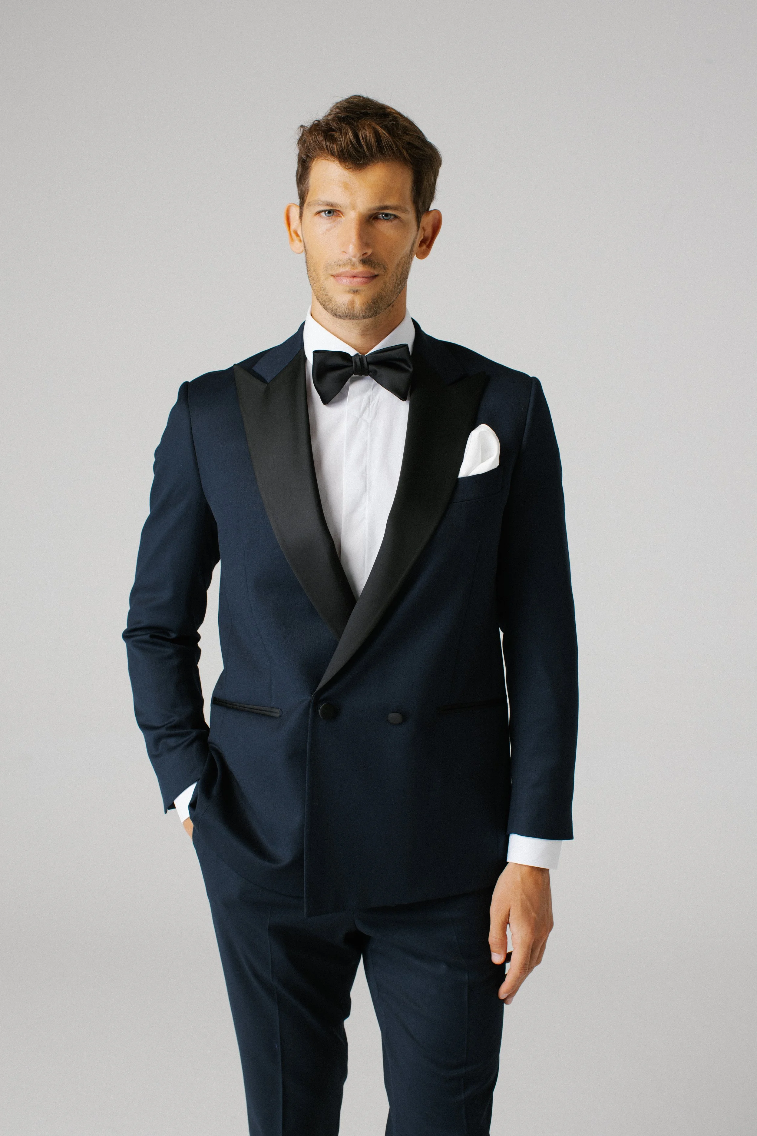 Uomo che indossa uno smoking blu con papillon nero e taschino bianco, sfondo bianco.