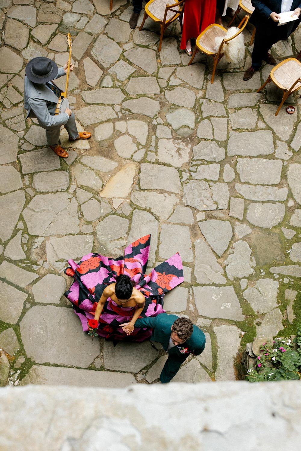 Una coppia si sposa, con la donna che indossa un abito nero con motivi rosa e rosso, e l'uomo in abiti eleganti, si tiene per mano sul pavimento di pietra. Sul pavimento sono presenti anche alcune sedie e un gruppo di persone dalle carine rigorose, u