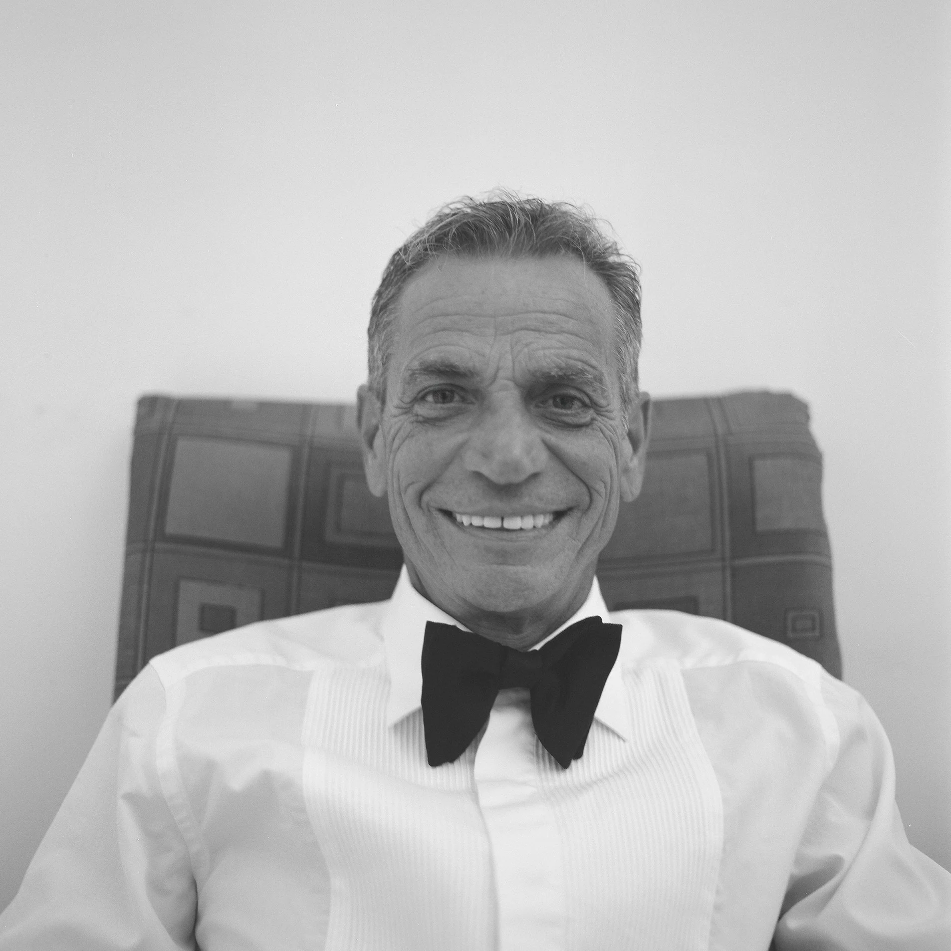 Uomo sorridente in abito bianco e papillon nero, seduto su una sedia con schienale decorato geometricamente.