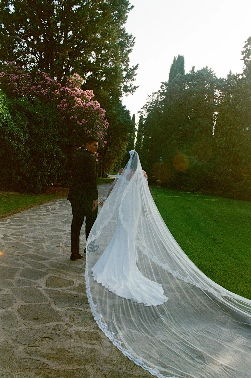 Una coppia di sposi, il marito in smoking e la sposa con un abito da matrimonio lungo con un velo esteso, camminano lungo un sentiero in un giardino con alberi e fiori, durante il tramonto.