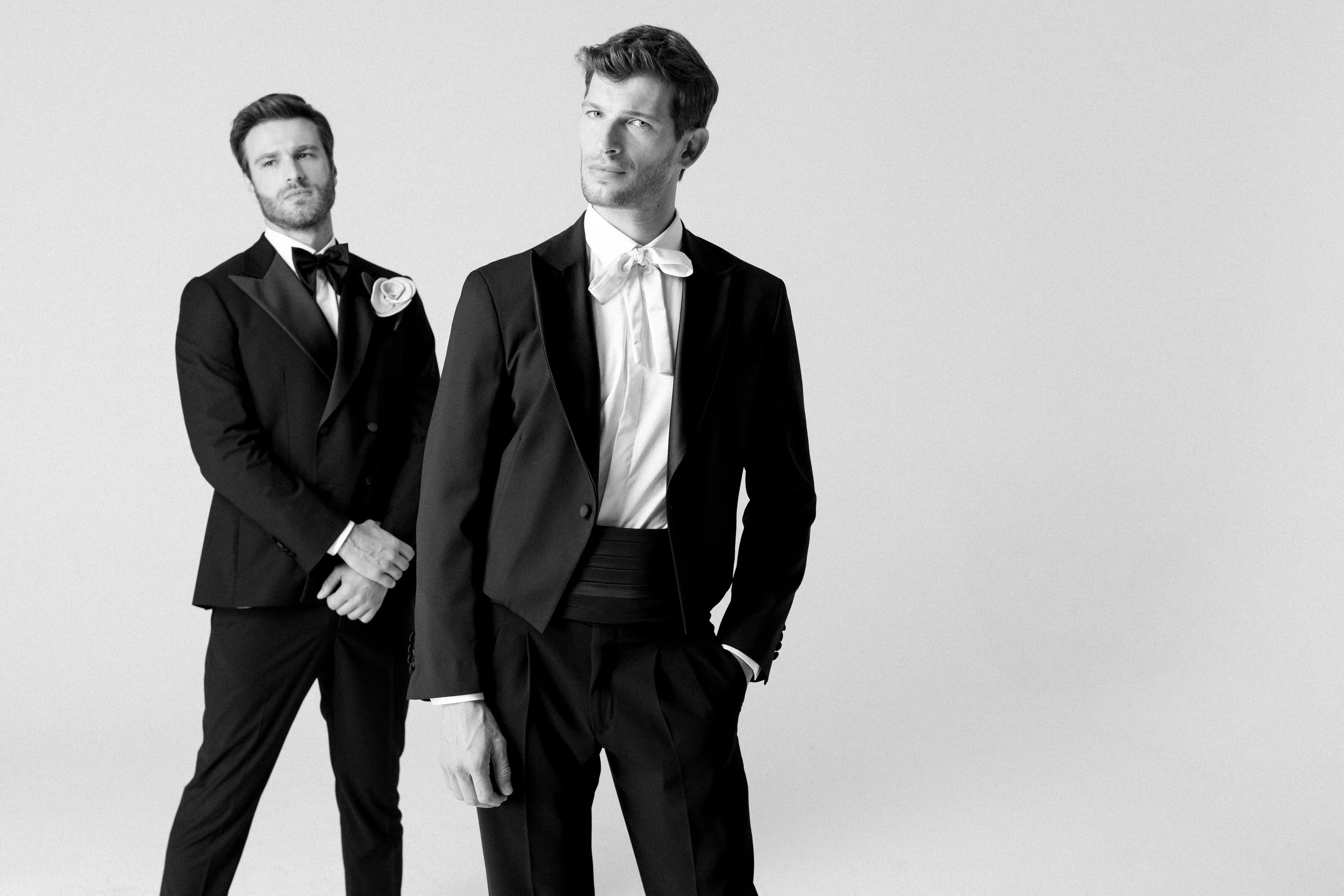 Due uomini vestiti elegantemente in stile formale, uno davanti e uno dietro, entrambi con abiti neri e camicie bianche, uno con papillon e l'altro con cravatta, in un ambiente bianco