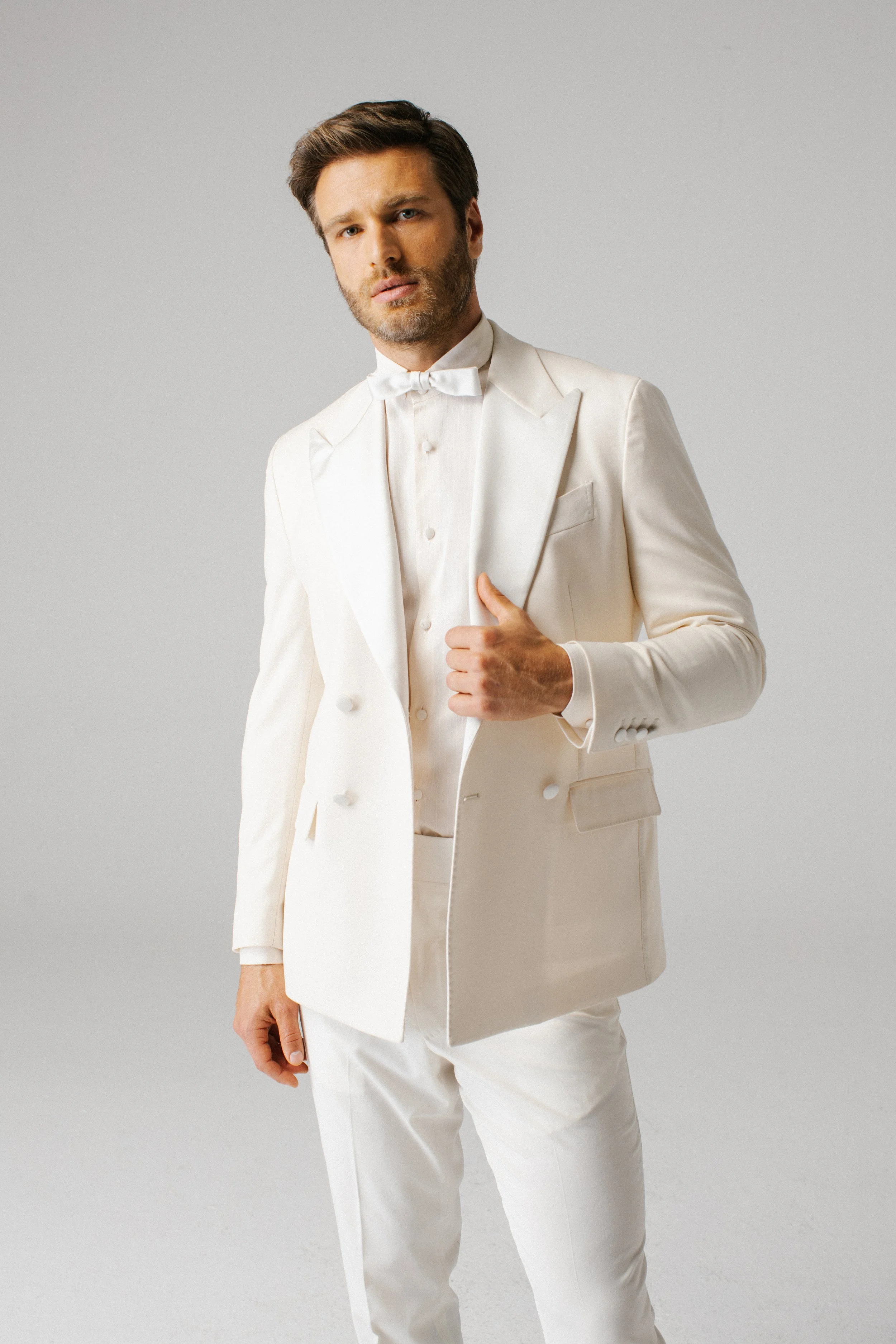 Uomo con abiti da sera di colore bianco, giacca doppio petto, camicia e papillon, in posa contro sfondo grigio chiaro.