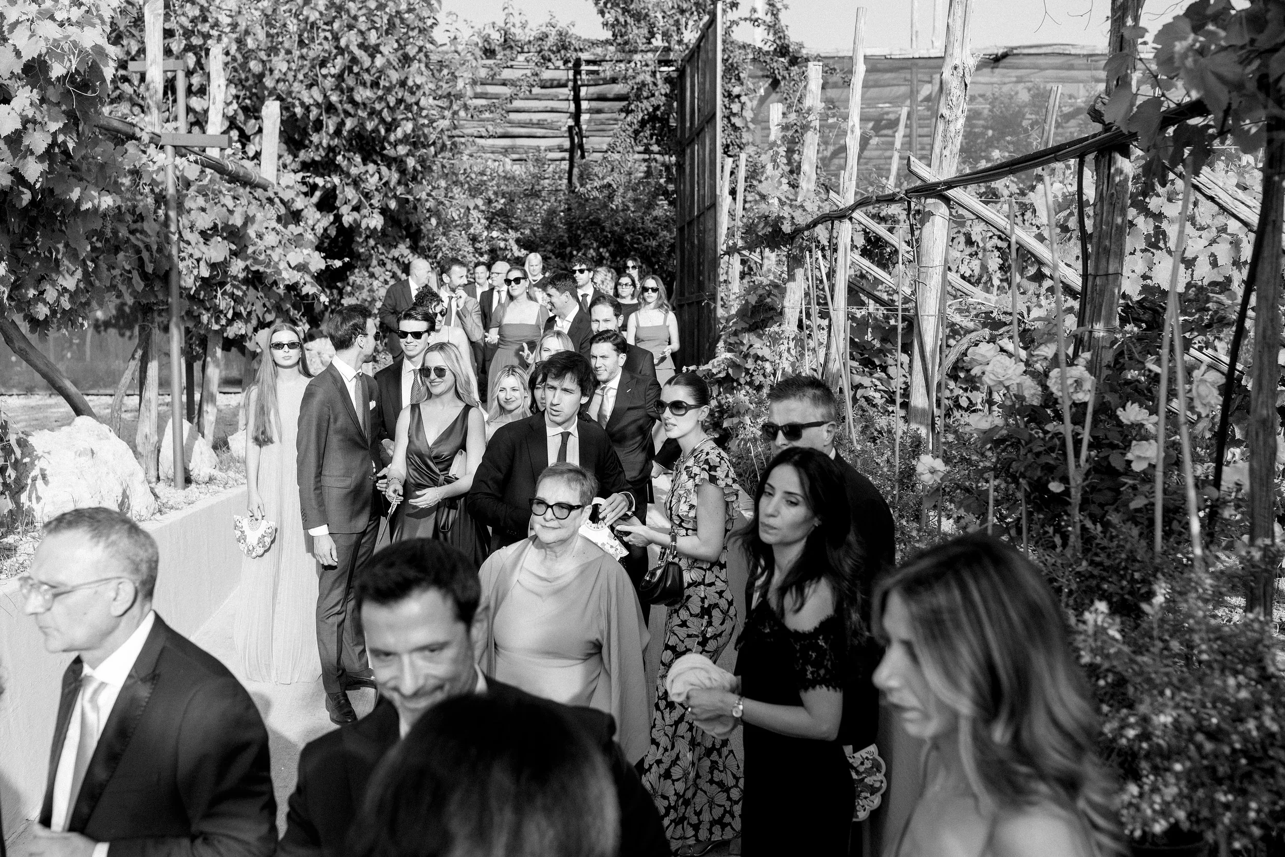 Una fila di persone eleganti in un giardino con piante e viti, alcune indossano occhiali da sole, sembrano in attesa di entrare o partecipare a un evento all'aperto.