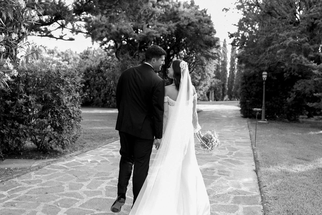 Una coppia di sposi in abiti da matrimonio si tiene per mano mentre camminano lungo un sentiero in un parco, circondati da alberi e vegetazione, in una scena di matrimonio romantica in bianco e nero.