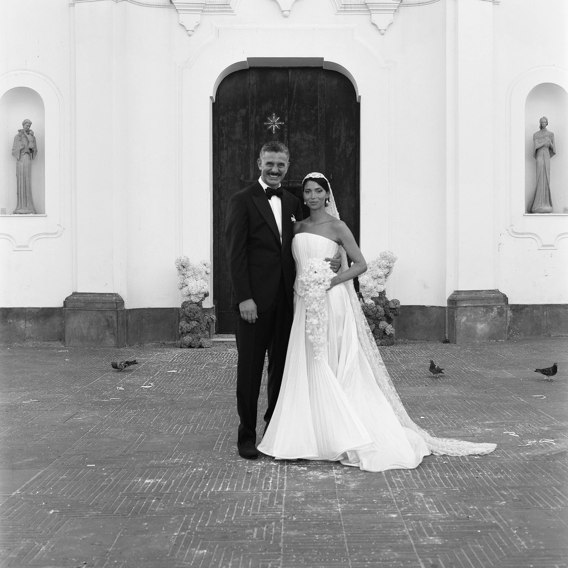 Una coppia di sposi in abiti da matrimonio davanti a una chiesa, in bianco e nero.