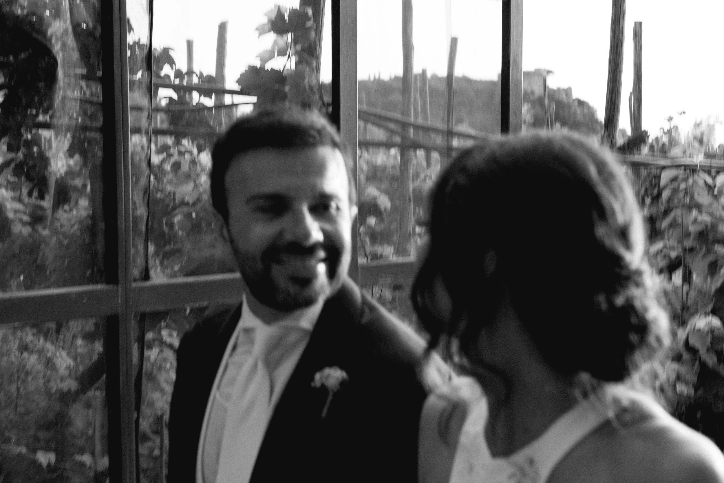 Una coppia in abiti eleganti si abbraccia in un ambiente naturale, con una cornice di piante e un paesaggio in lontananza.