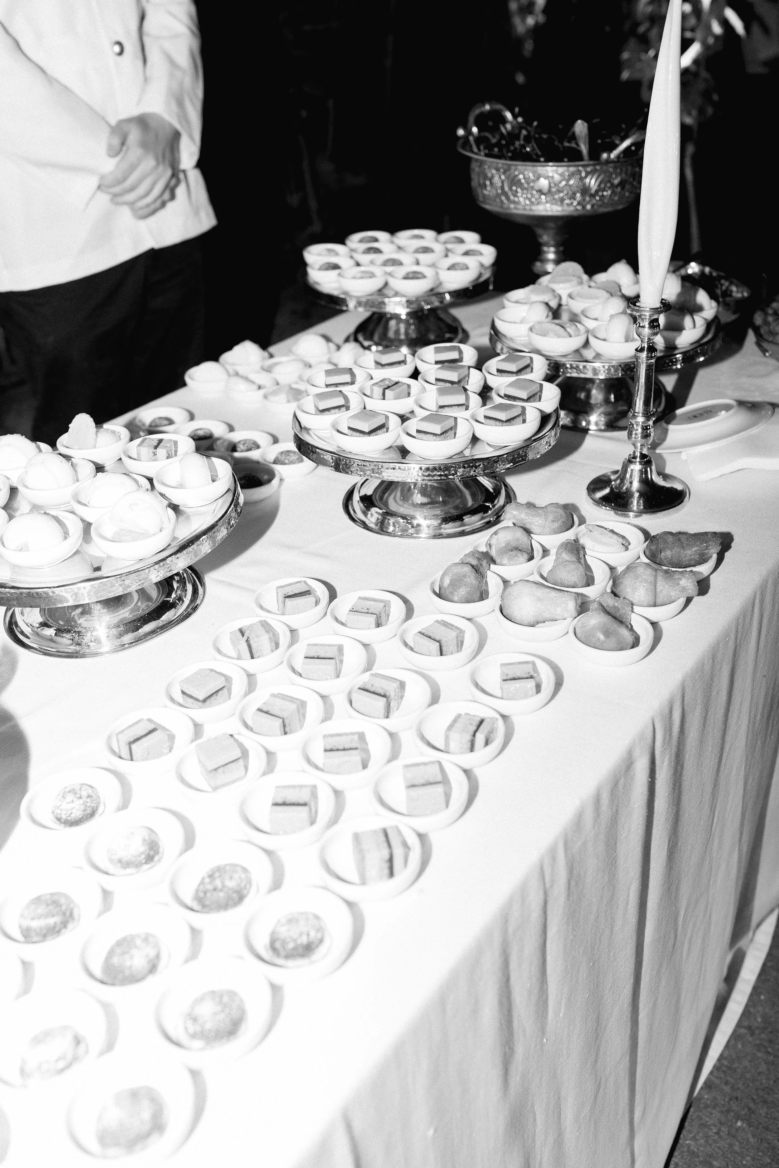 Tavolo con vari dessert e dolci disposti su vassoi e piattini, con un commesso in piedi in background.