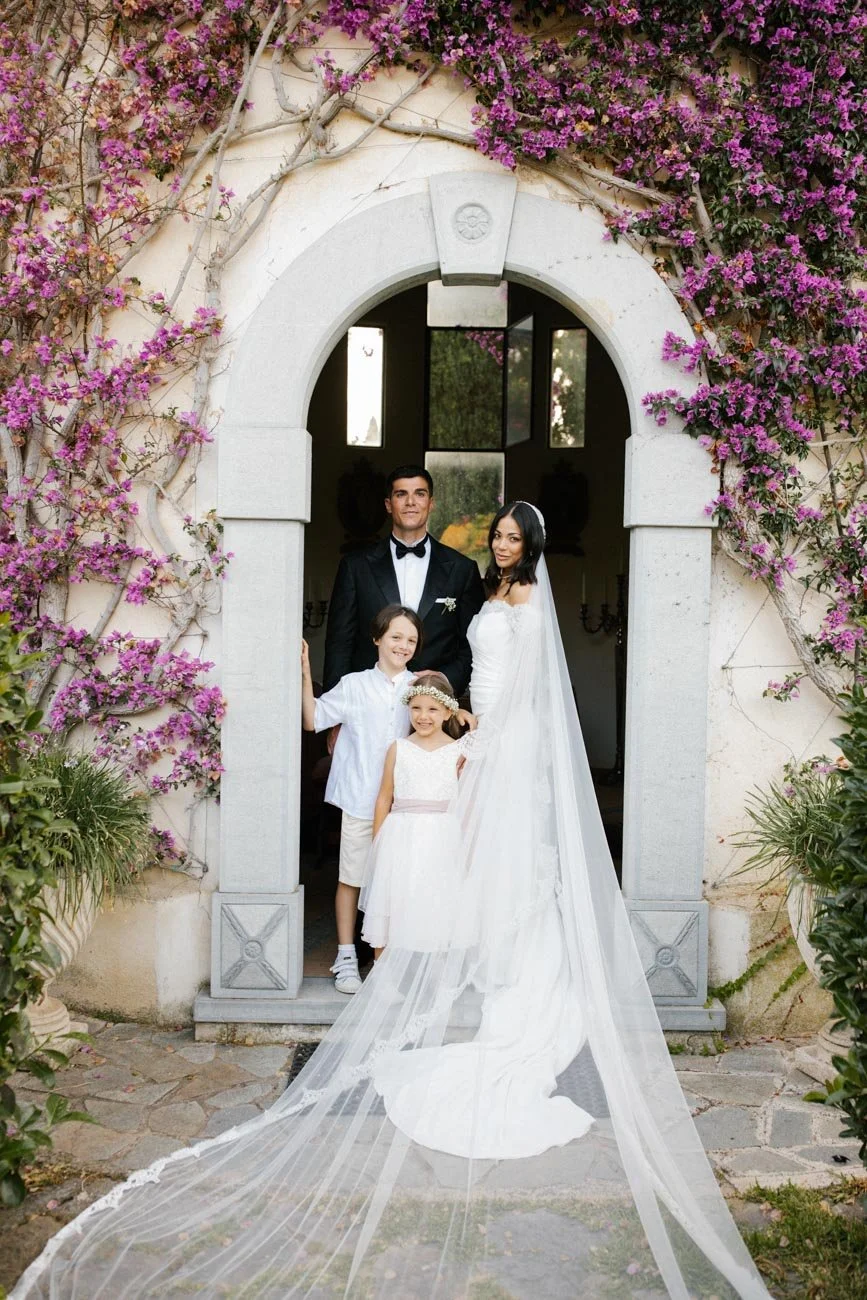 Una coppia di sposi insieme a due bambini sorridenti in piedi sotto un arco decorato con fiori viola, davanti a una chiesa o un edificio storico con pareti chiare.