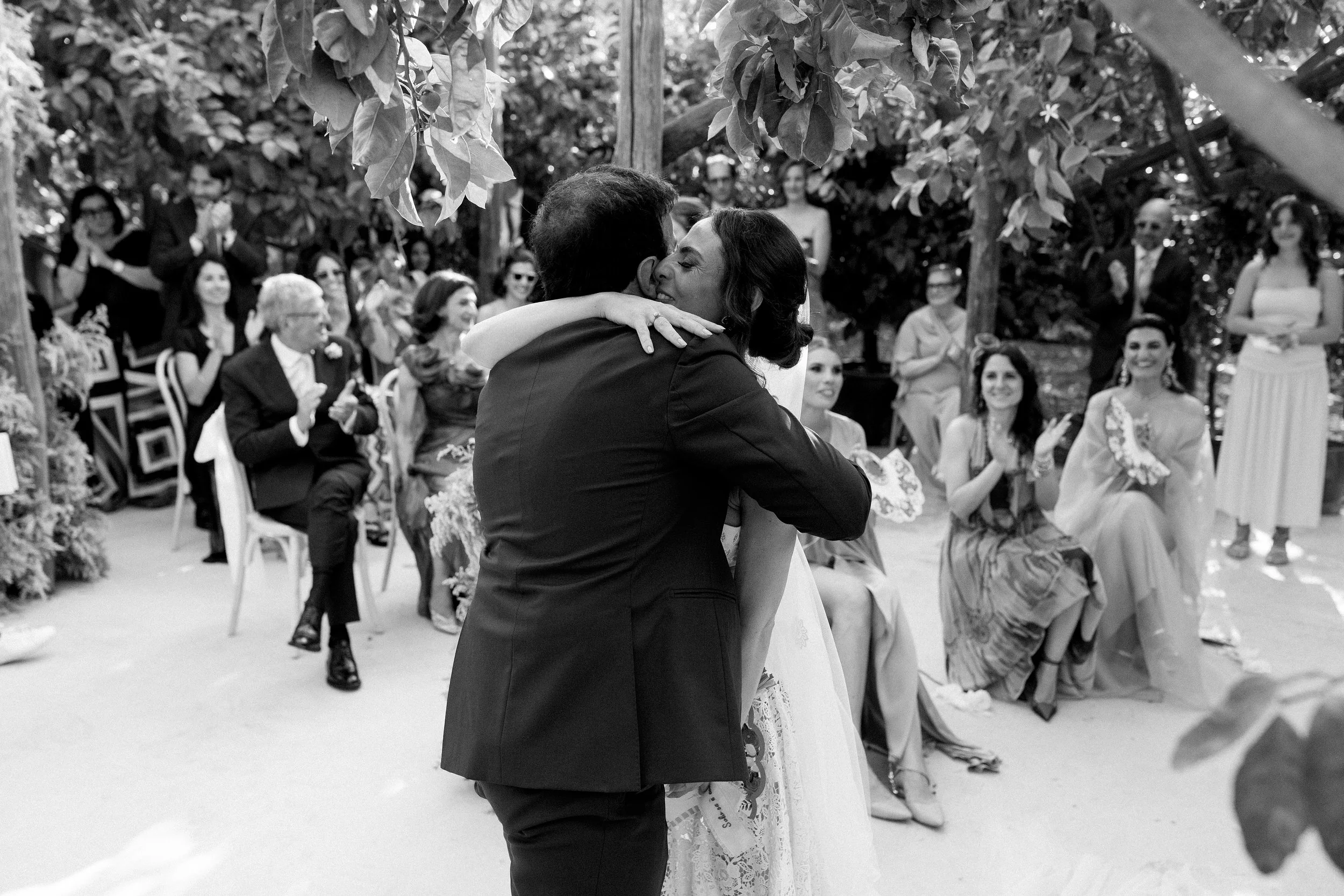 Un matrimonio si abbraccia durante la cerimonia con invitati seduti e che applaudono, in un ambiente all'aperto circondato da alberi.