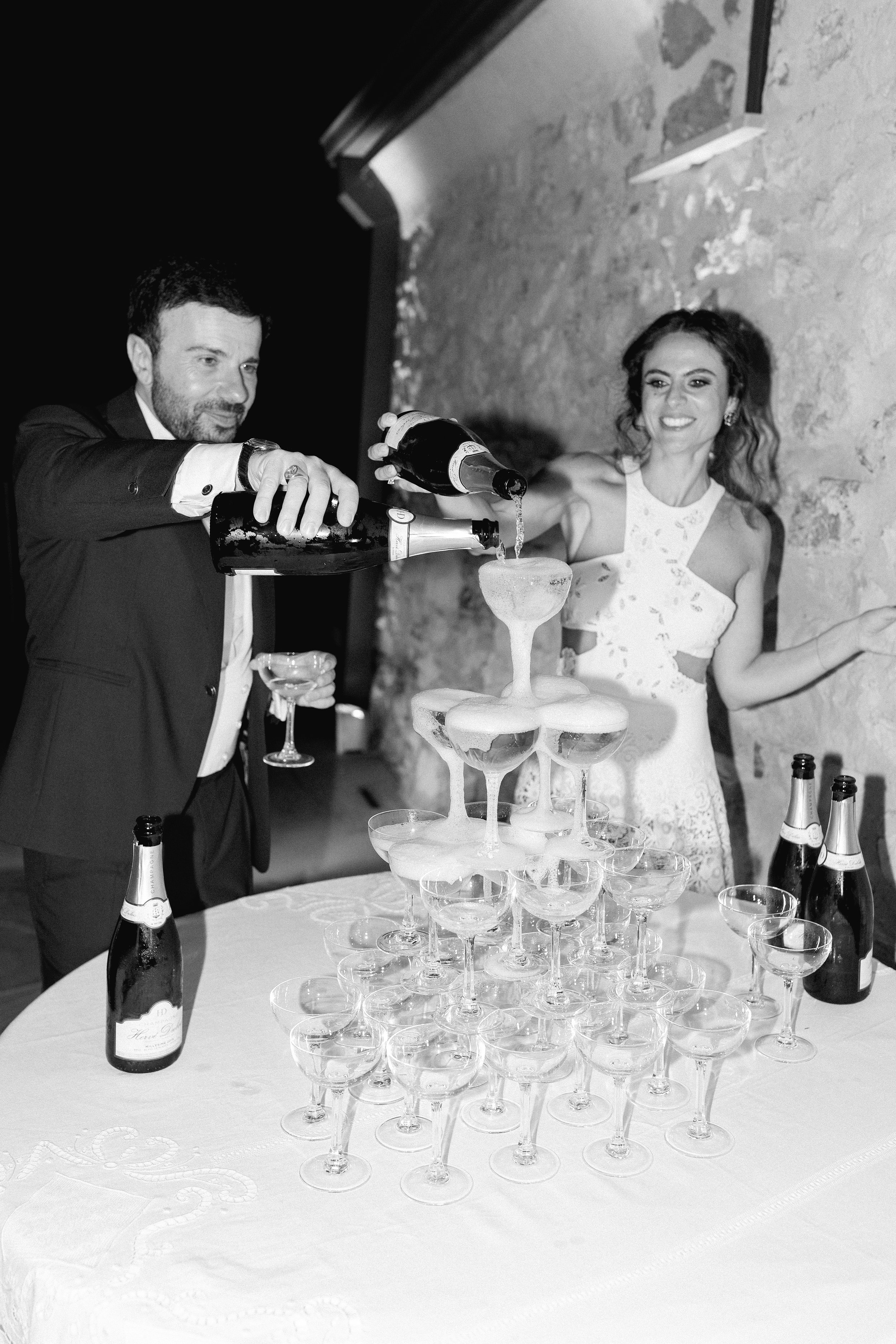Una coppia sorride mentre versa champagne in un capolavoro di champagne a forma di torre di bicchieri su un tavolo, in un ambiente elegante con pareti in pietra, decorato per una festa.
