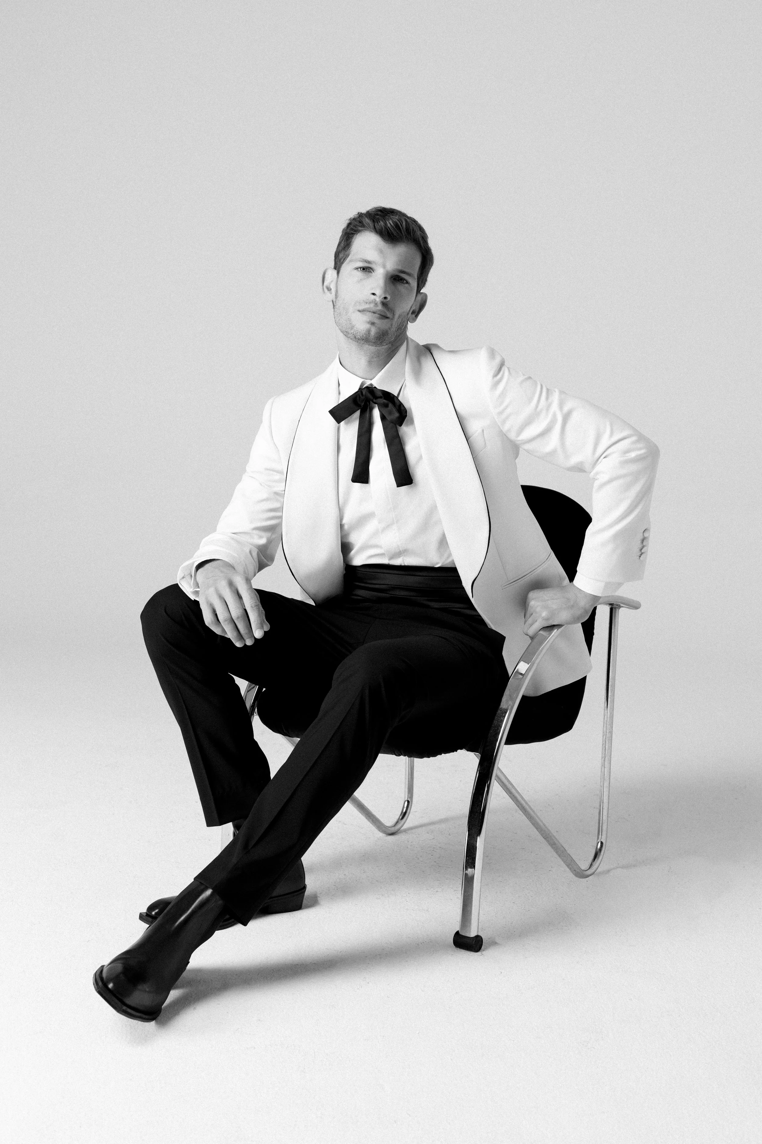 Uomo seduto su una sedia, indossa una giacca bianca con dettagli neri, una camicia bianca con un fiocco nero e pantaloni neri, ritratto in bianco e nero.