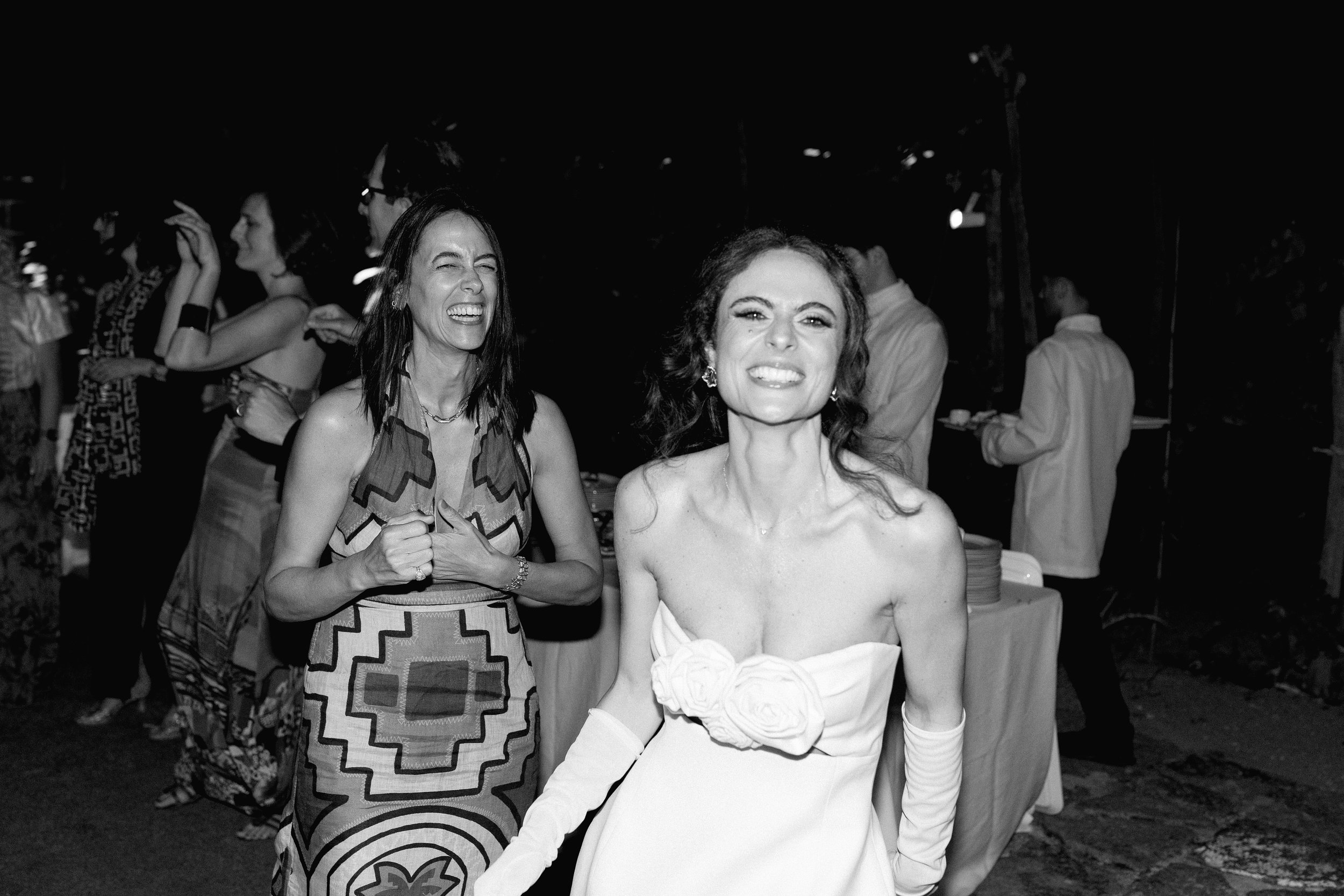 Due donne sorridenti vestite elegantemente alla festa, una indossa un abito bianco con rose e guanti lunghi, l'altra un vestito con un motivo geometrico, sullo sfondo altre persone al party.