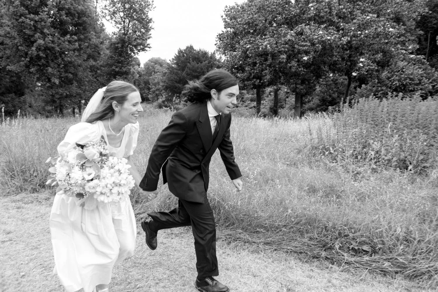 Una coppia vestita da sposi corretta nel prato con alberi sullo sfondo, sorridendo e che si tiene per mano.