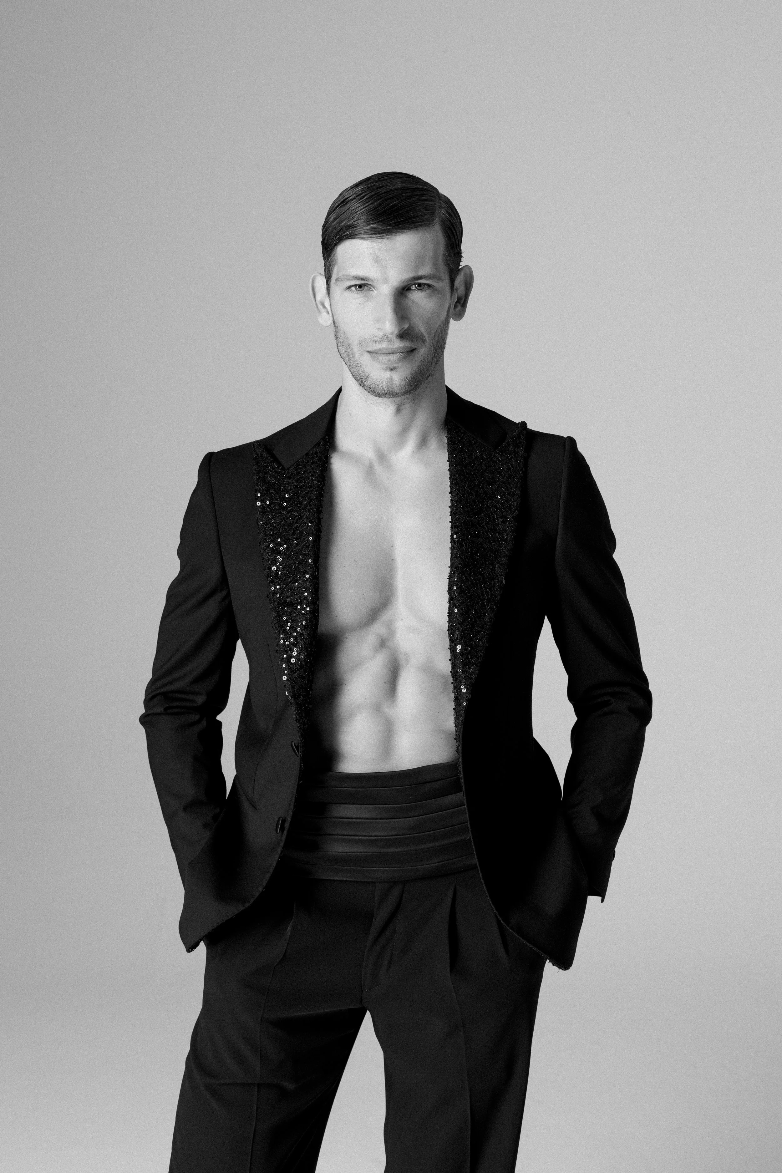 Uomo con giacca nera aperta e senza maglietta, in posa con le mani in tasca, in un'immagine in bianco e nero.