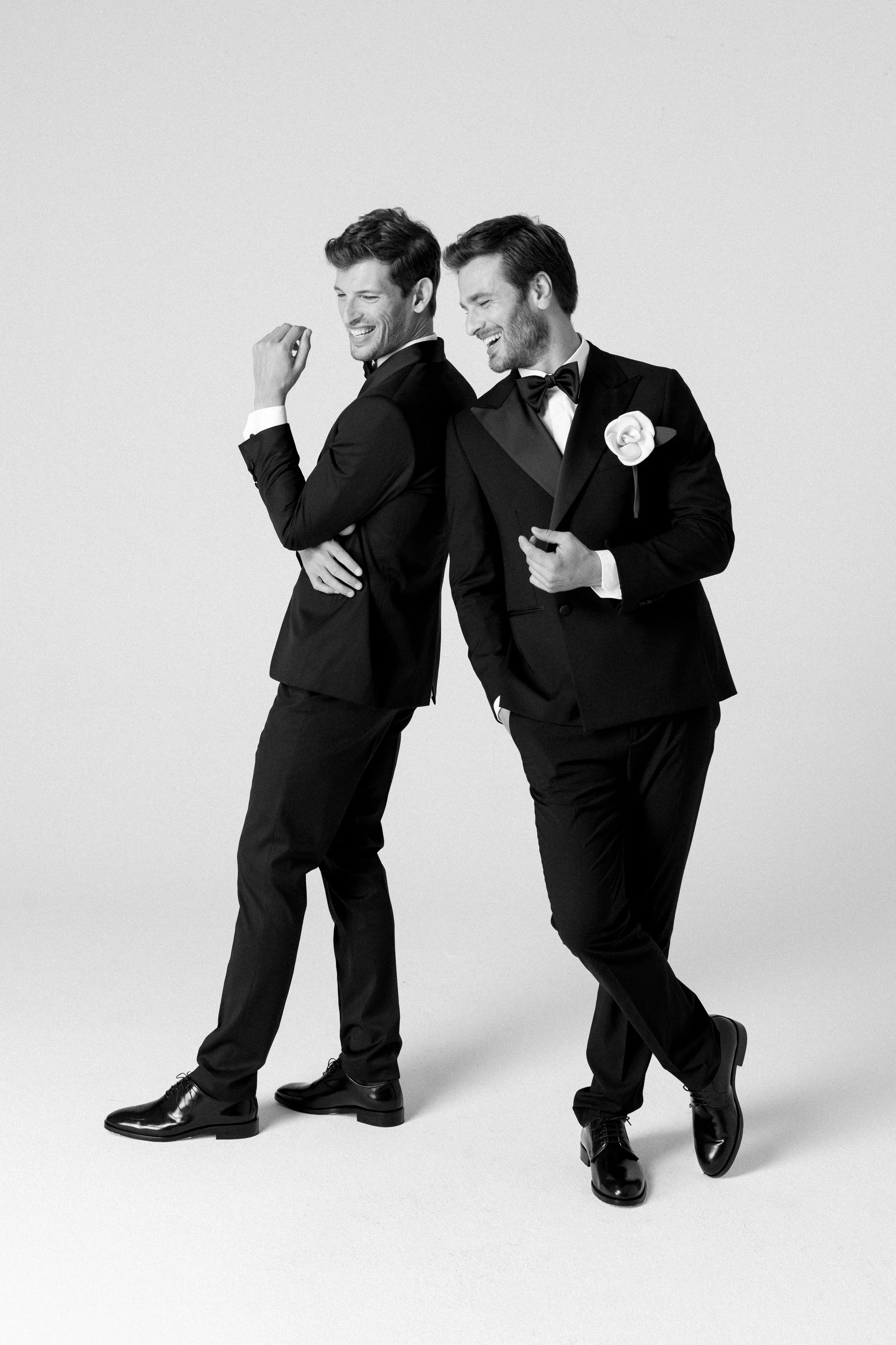 Due uomini eleganti in abiti formali sorridono e si divertono, uno tiene un fiore bianco in mano, sfondo bianco.