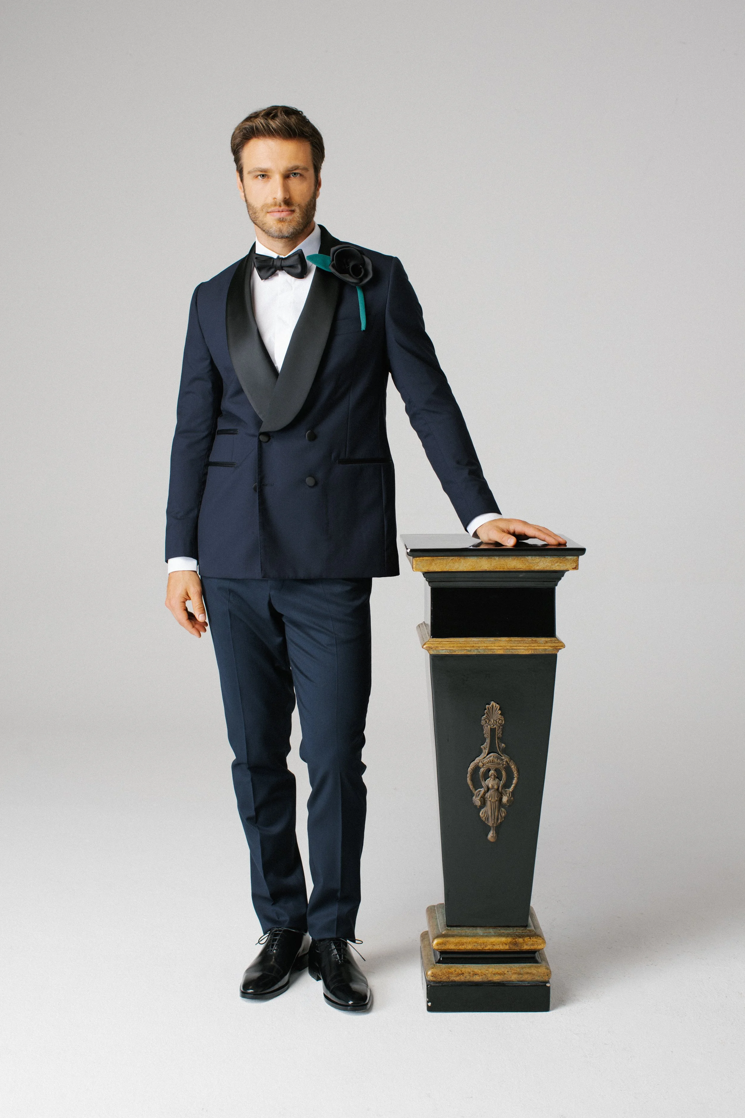 Uomo in abito da sera con fiocco, con fiore sulla spalla, appoggiato a un piedistallo con decorazioni d'oro e rilievo, sfondo neutro.