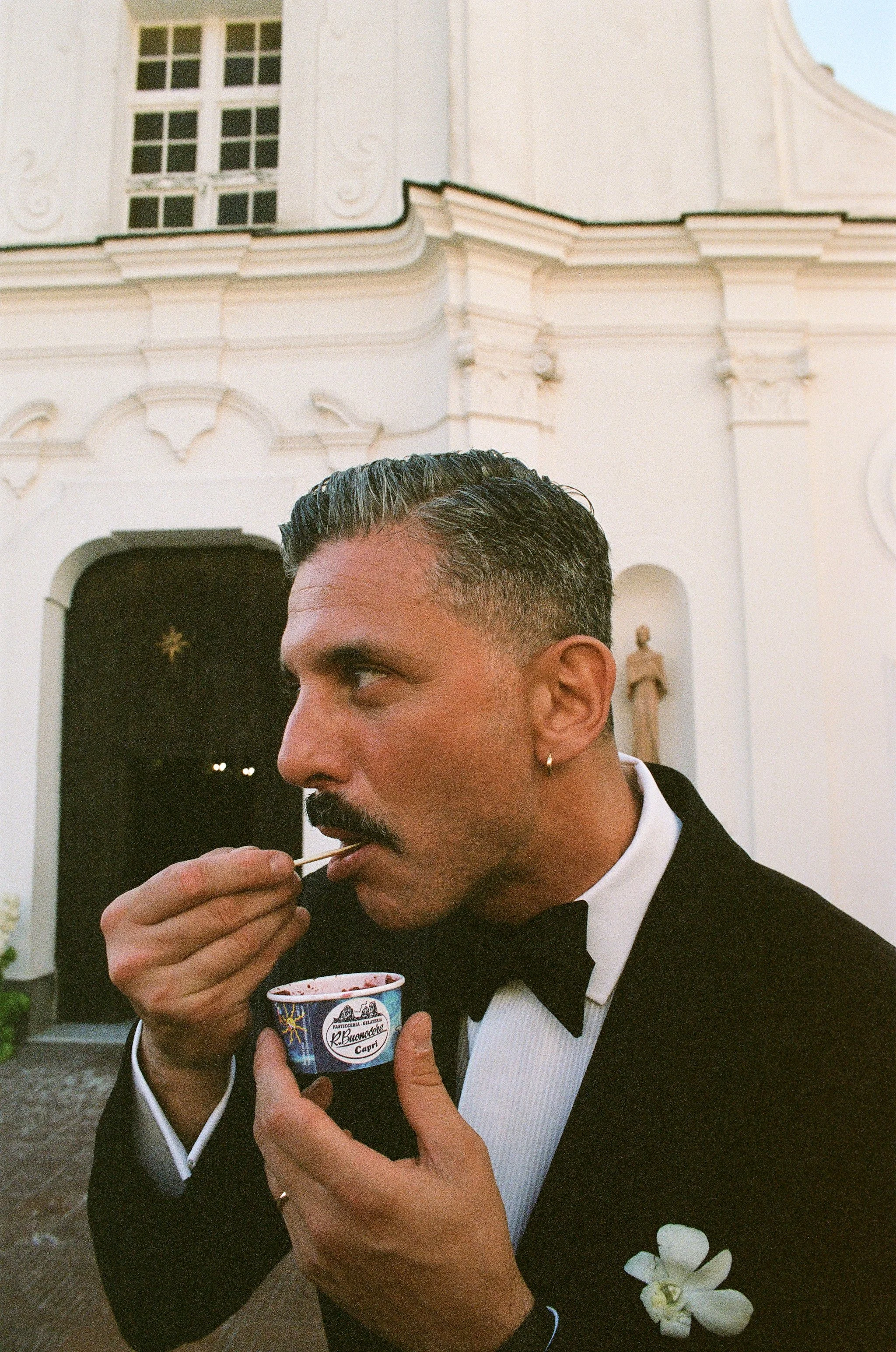 Uomo elegante che mangia un gelato fuori da una chiesa bianca, indossa un smoking con fiore bianco sulla giacca.